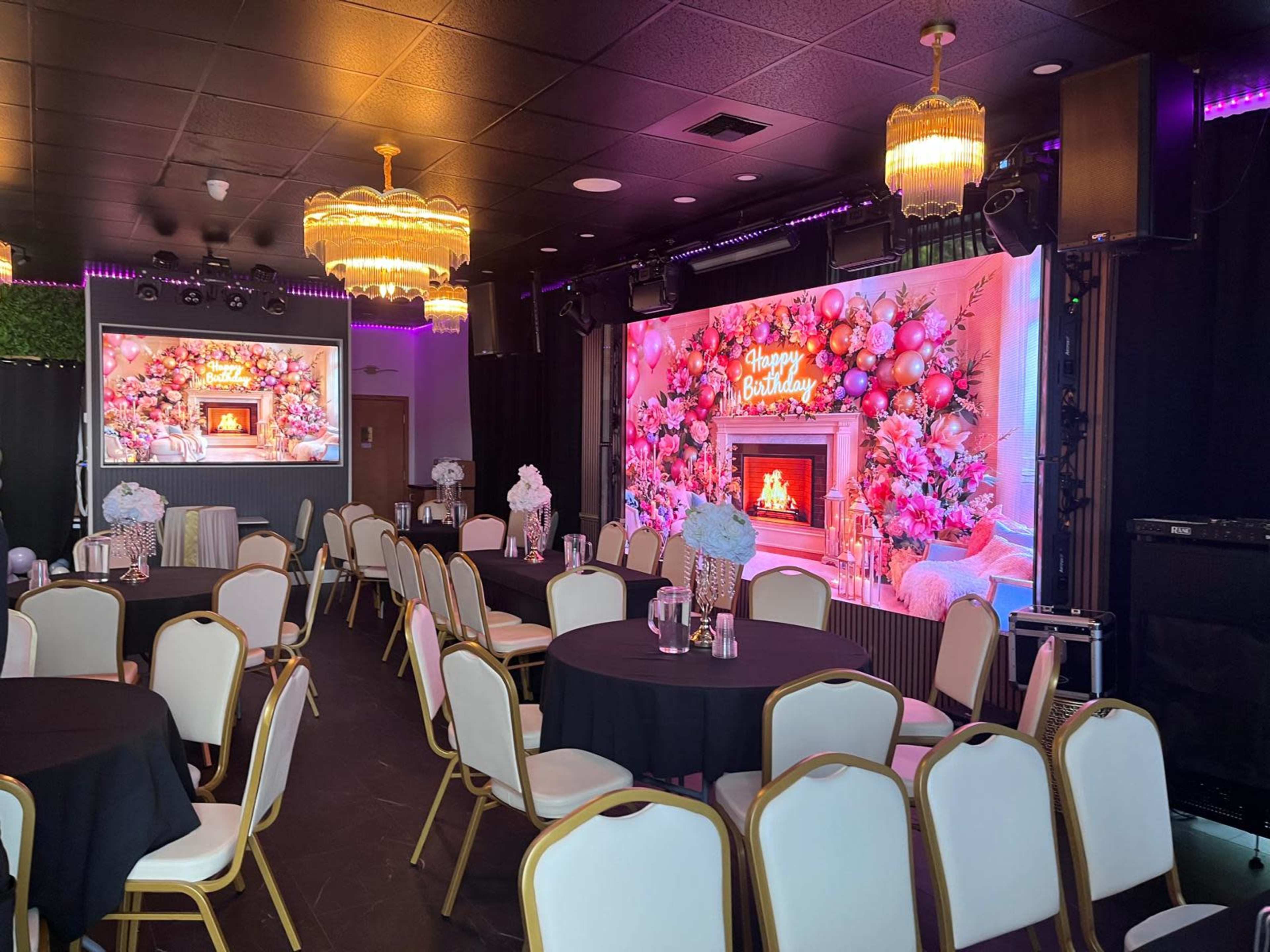 A decorated event space features round tables with black tablecloths and white chairs, highlighted by a vibrant birthday-themed backdrop with a fireplace surrounded by floral arrangements.