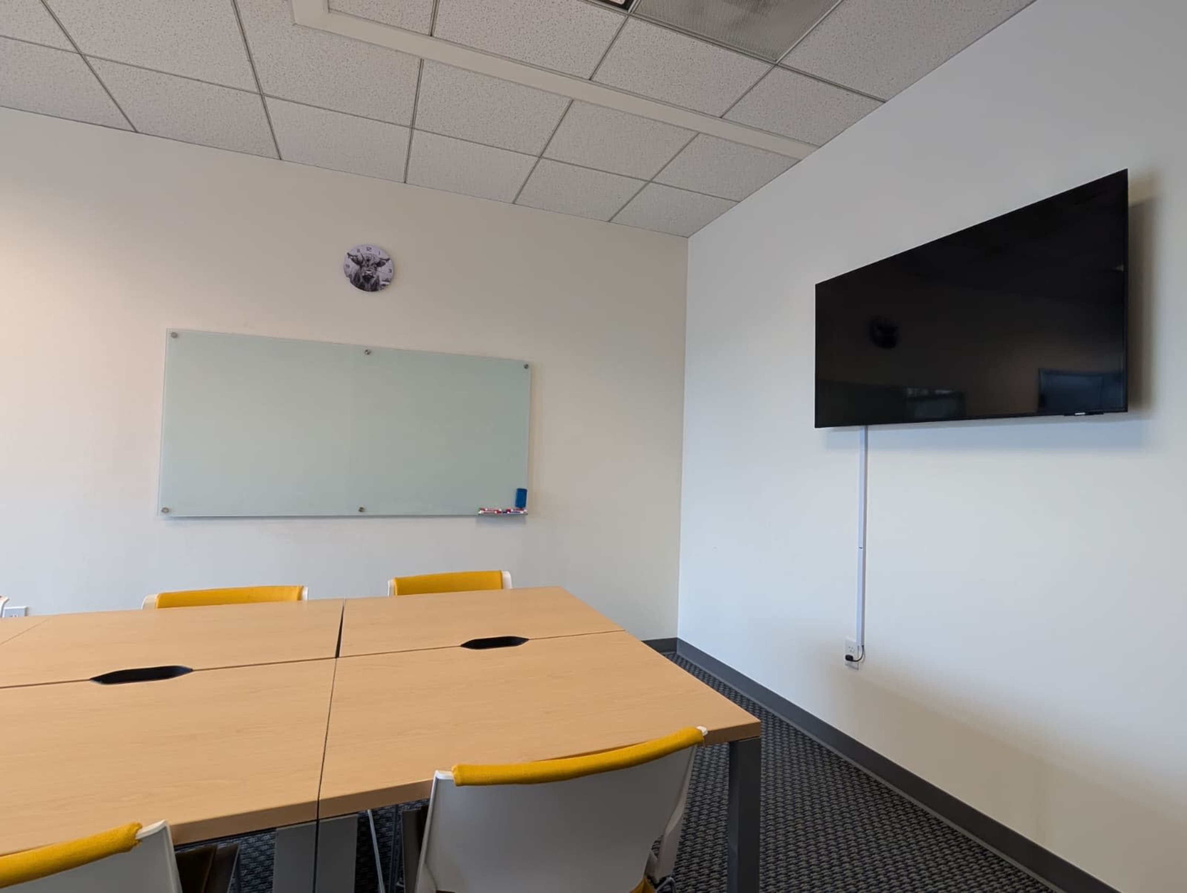 A conference room features a large table with four yellow chairs, a wall-mounted TV, a clock, and a glass whiteboard.