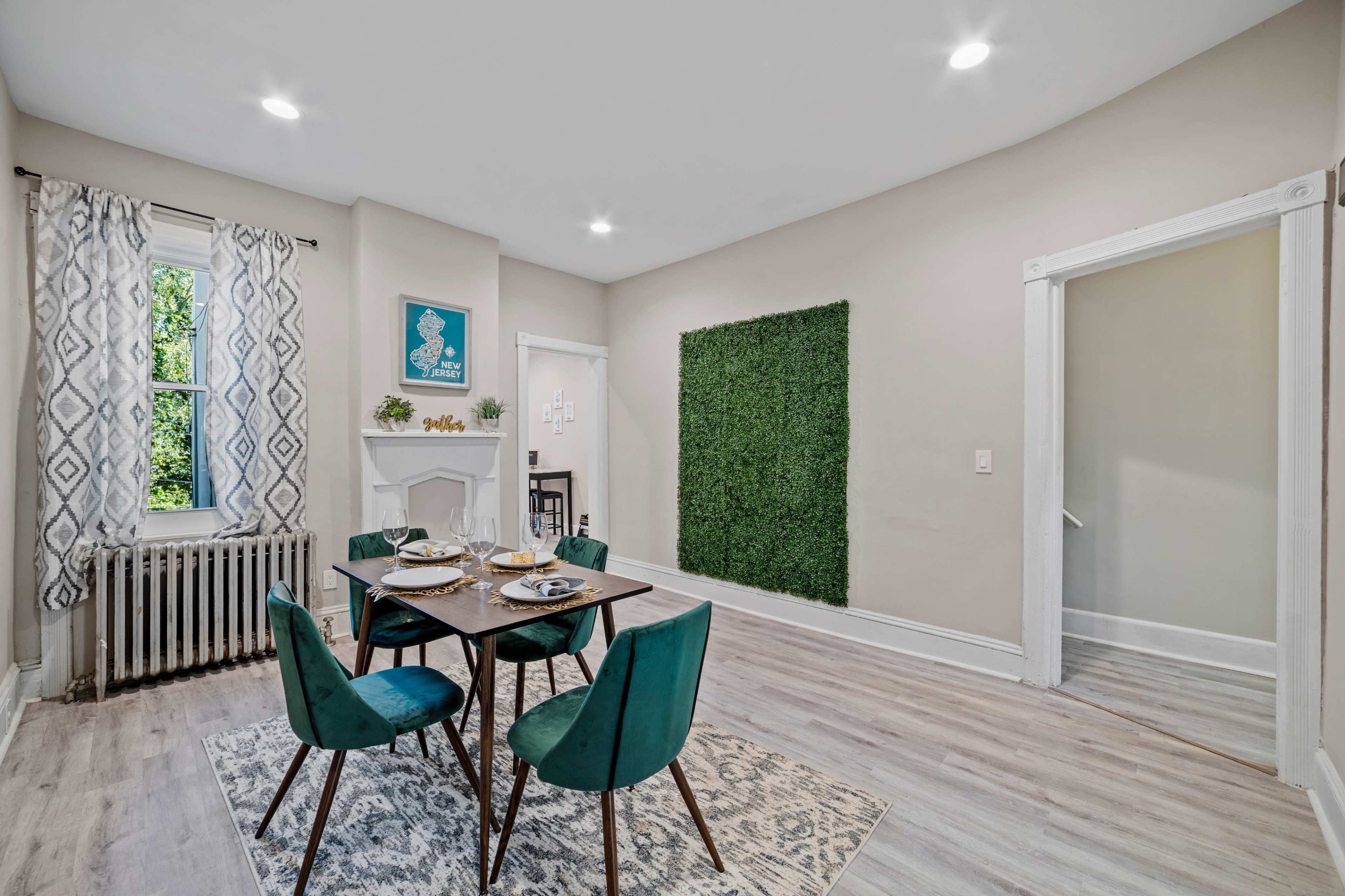 A dining area with a table set for four, surrounded by teal chairs, a window providing natural light, and a wall covered in greenery.