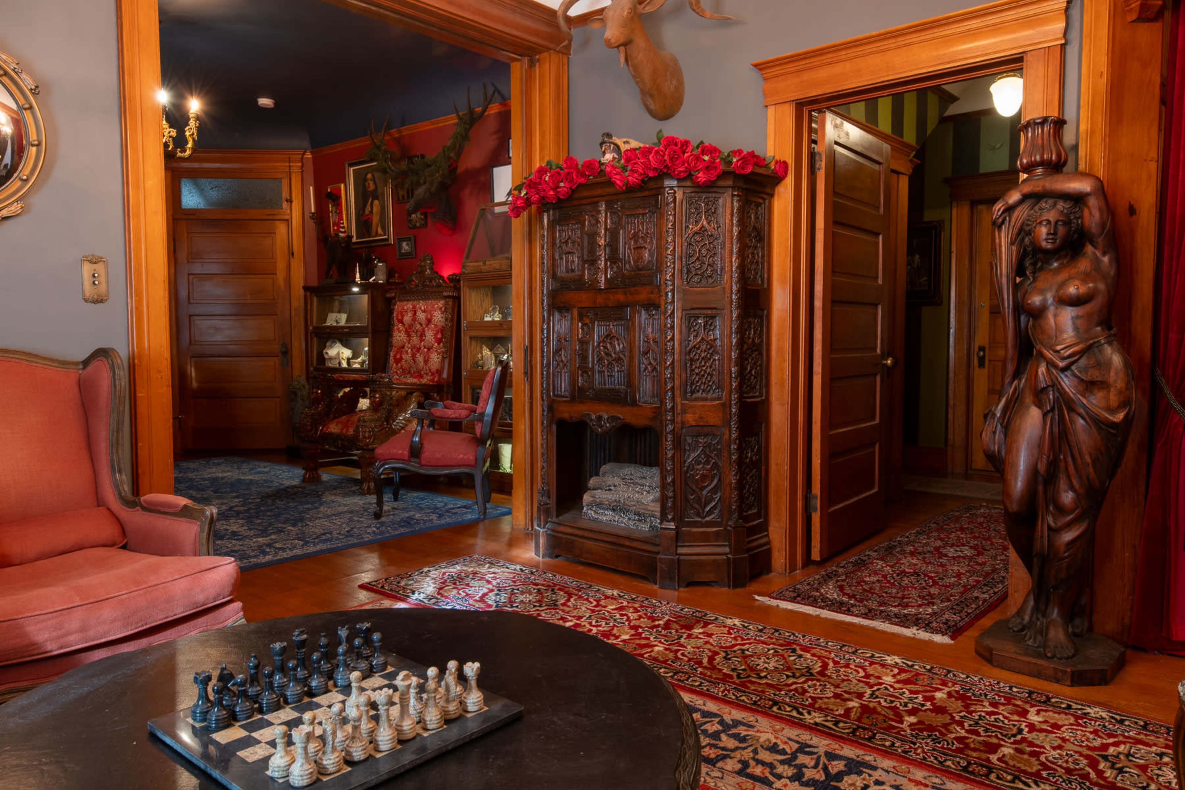 An ornate room featuring a large wooden chess set on a low table, a carved wooden statue of a woman, and decorative furniture, all surrounded by richly colored walls and rugs.
