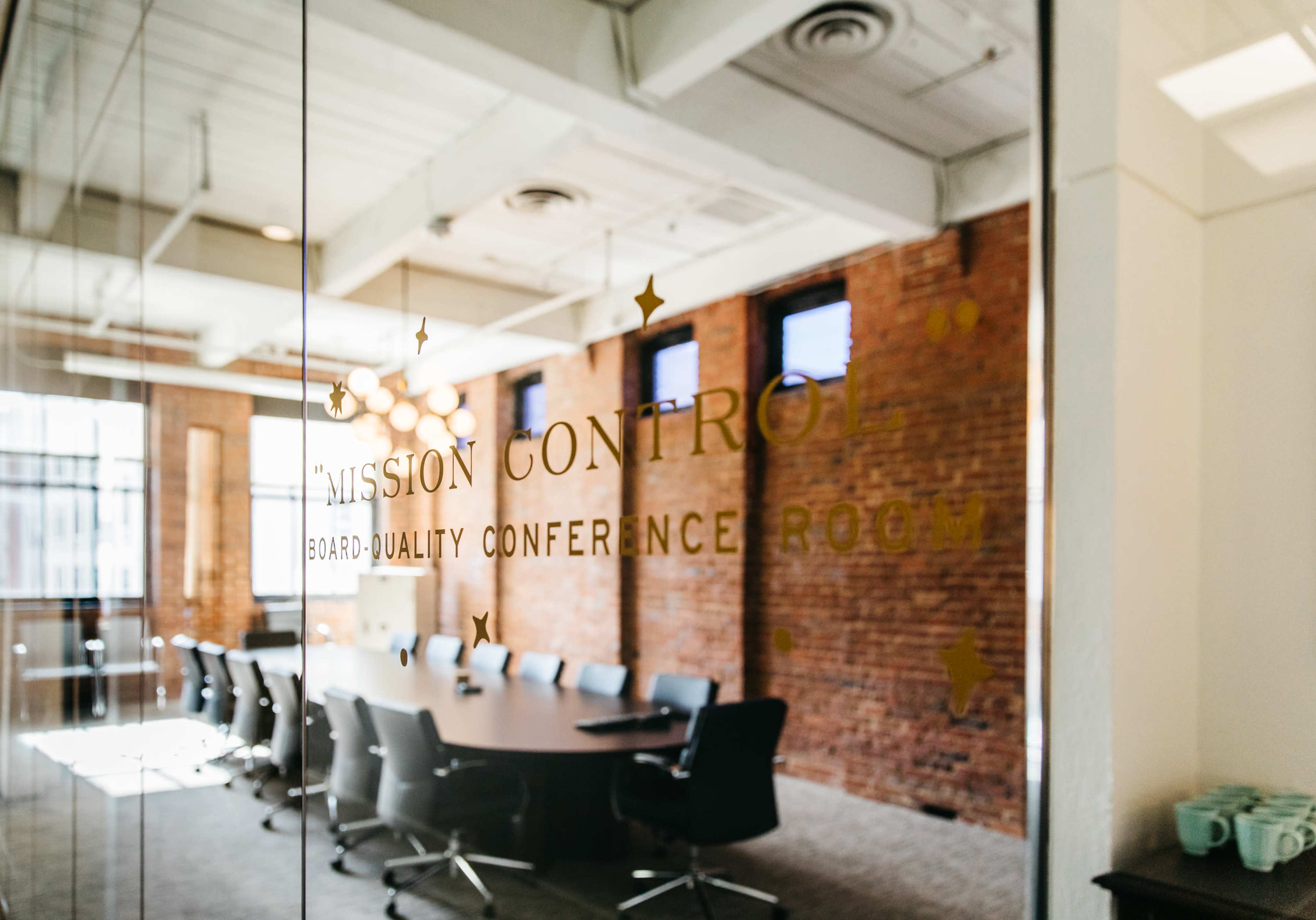 The image shows a conference room with a large round table and dark chairs, featuring exposed brick walls and large windows.