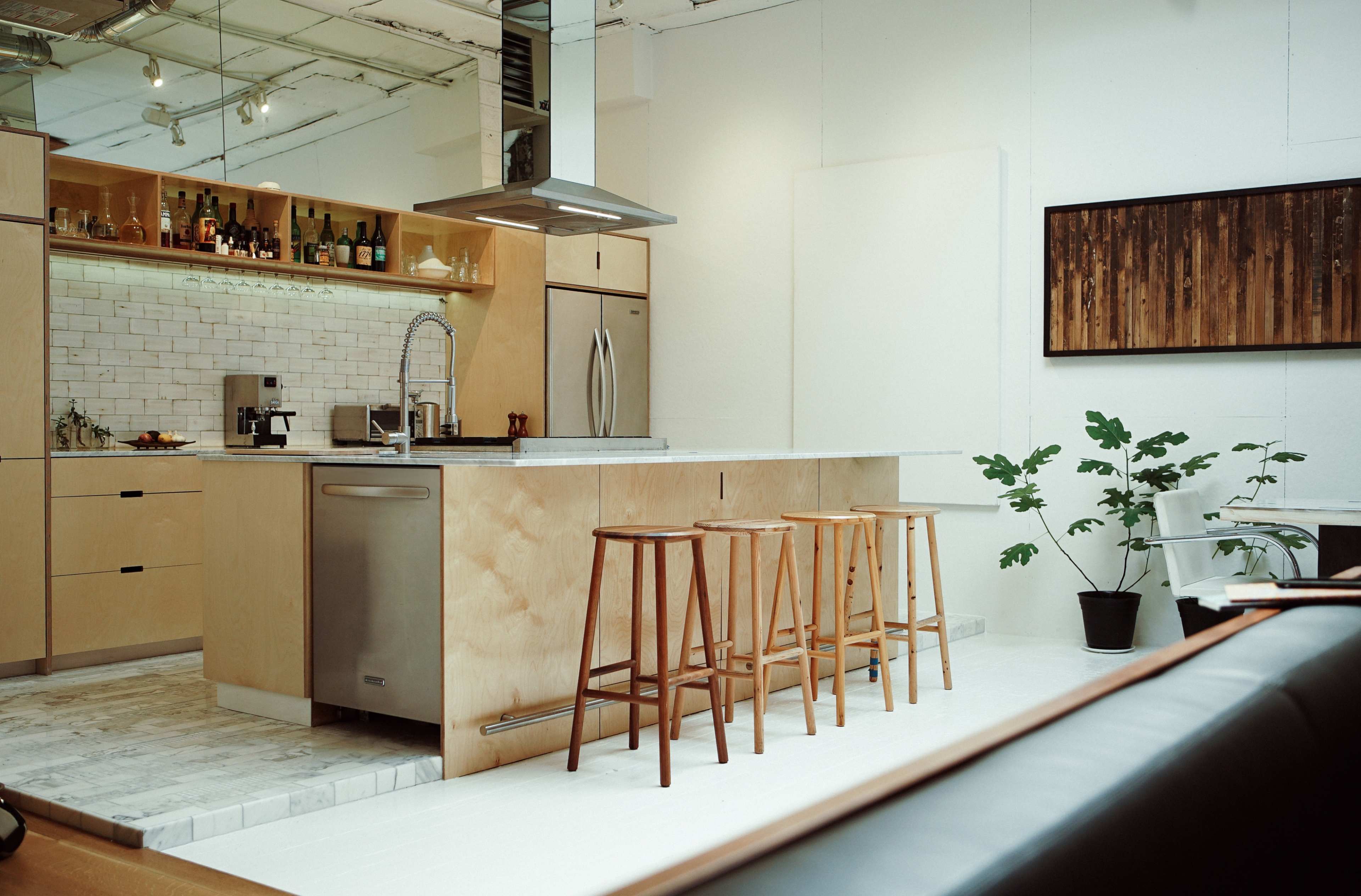 A modern kitchen featuring a central island with three wooden stools, stainless steel appliances, and a white tiled backsplash.