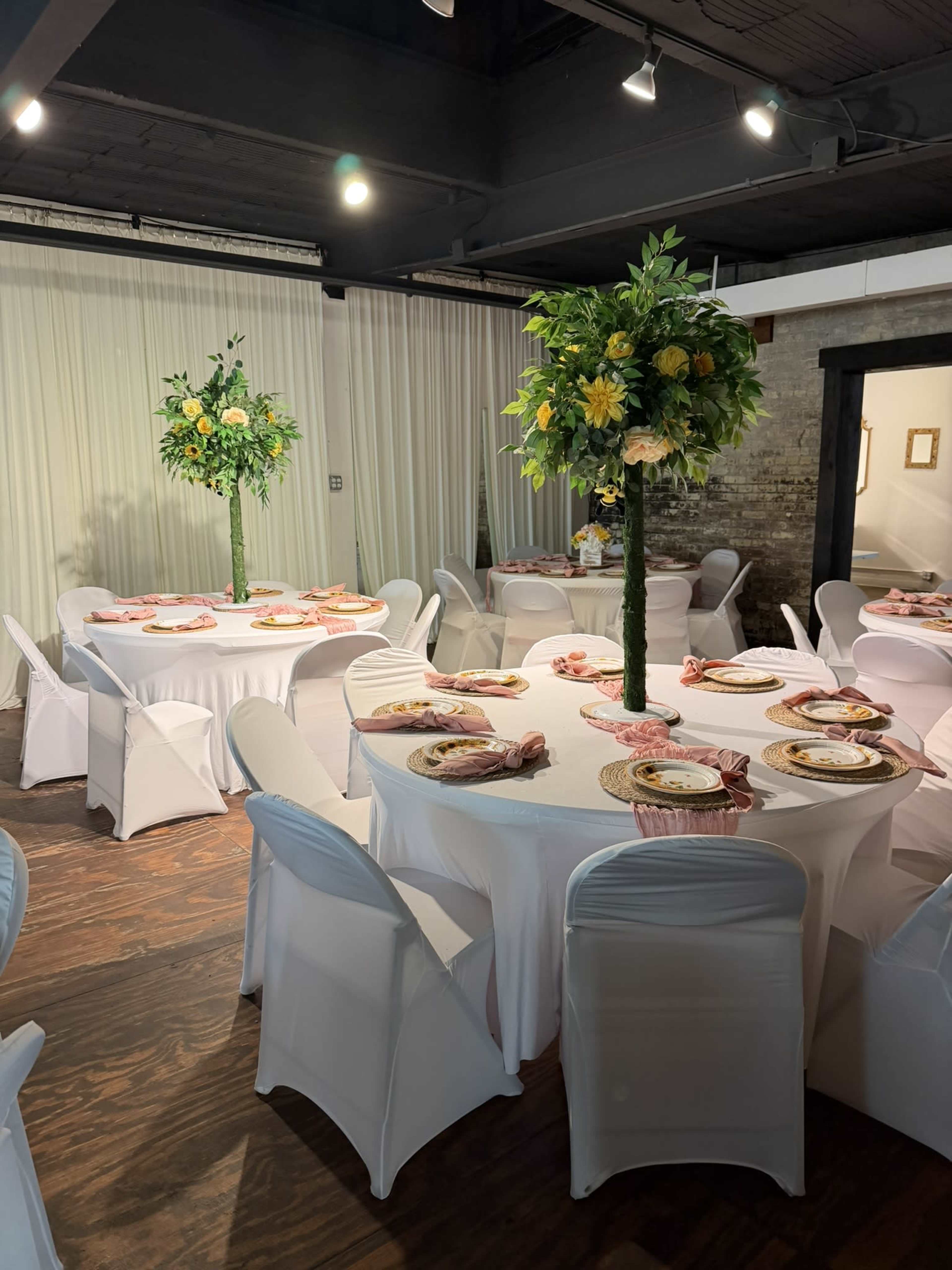 The image shows a banquet area with round tables covered in white tablecloths, each adorned with floral centerpieces and neatly set plates and napkins.