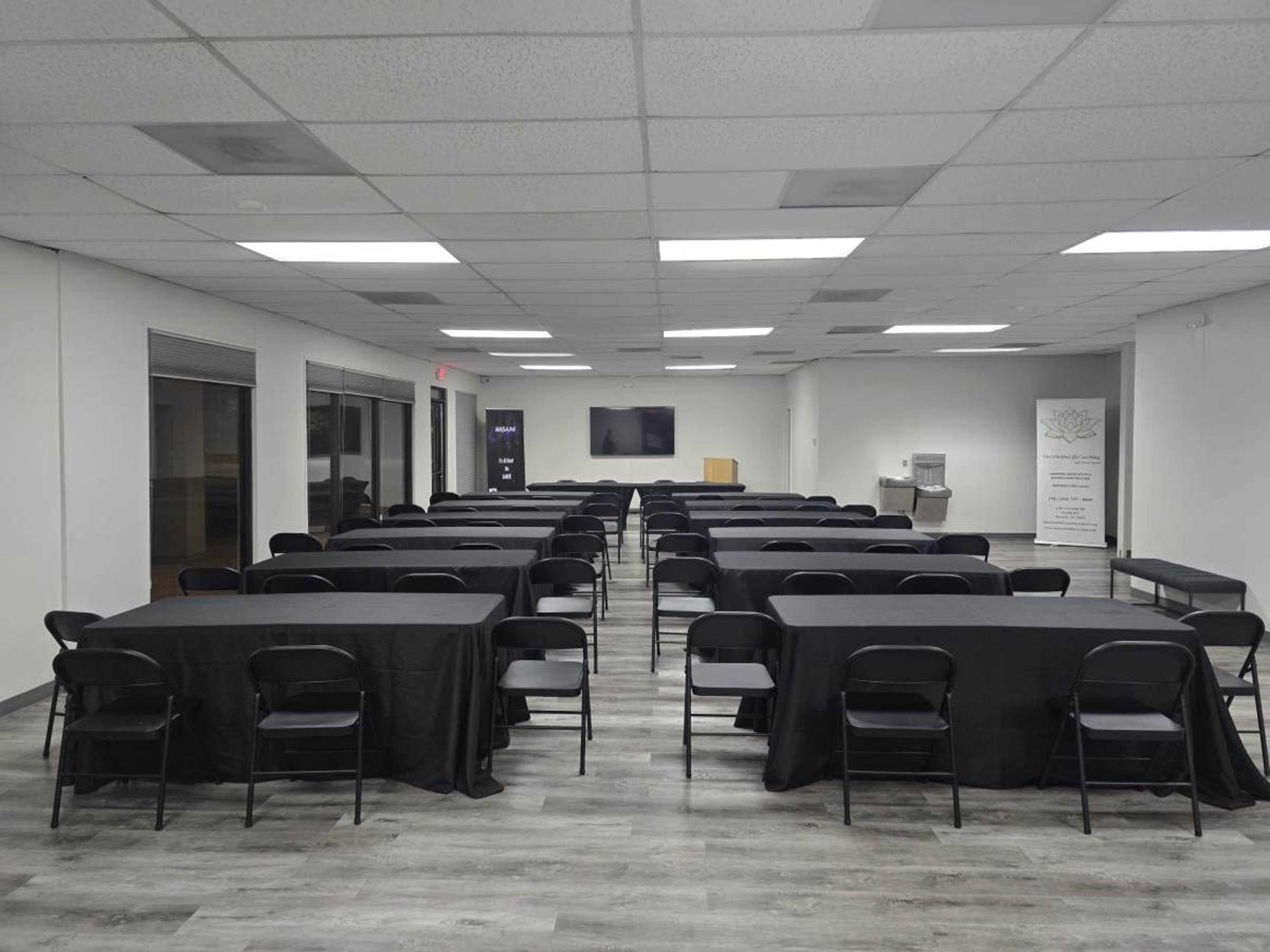 A large, empty room is arranged with multiple rows of black tables and chairs, featuring a presentation screen at one end.