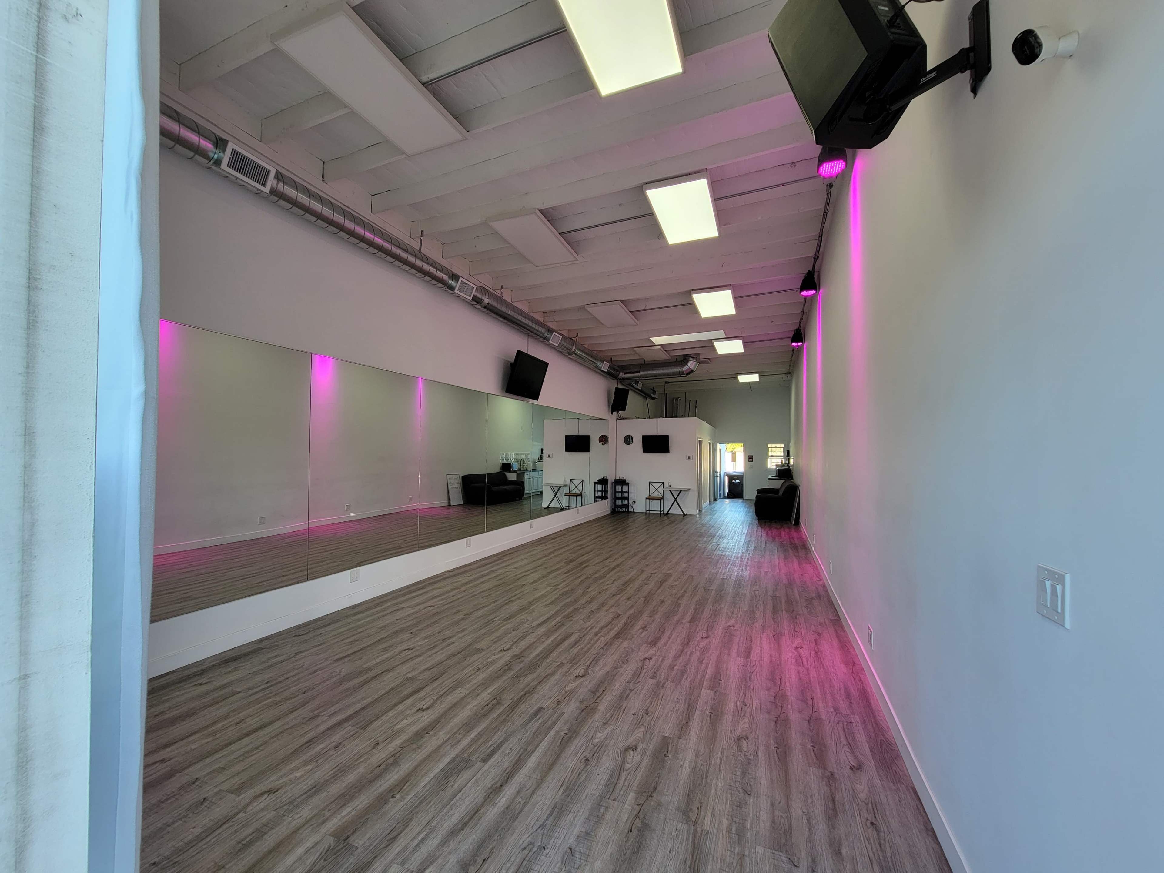 The image shows a spacious, empty studio with light wood flooring, white walls, and pink LED lighting along the mirrors.