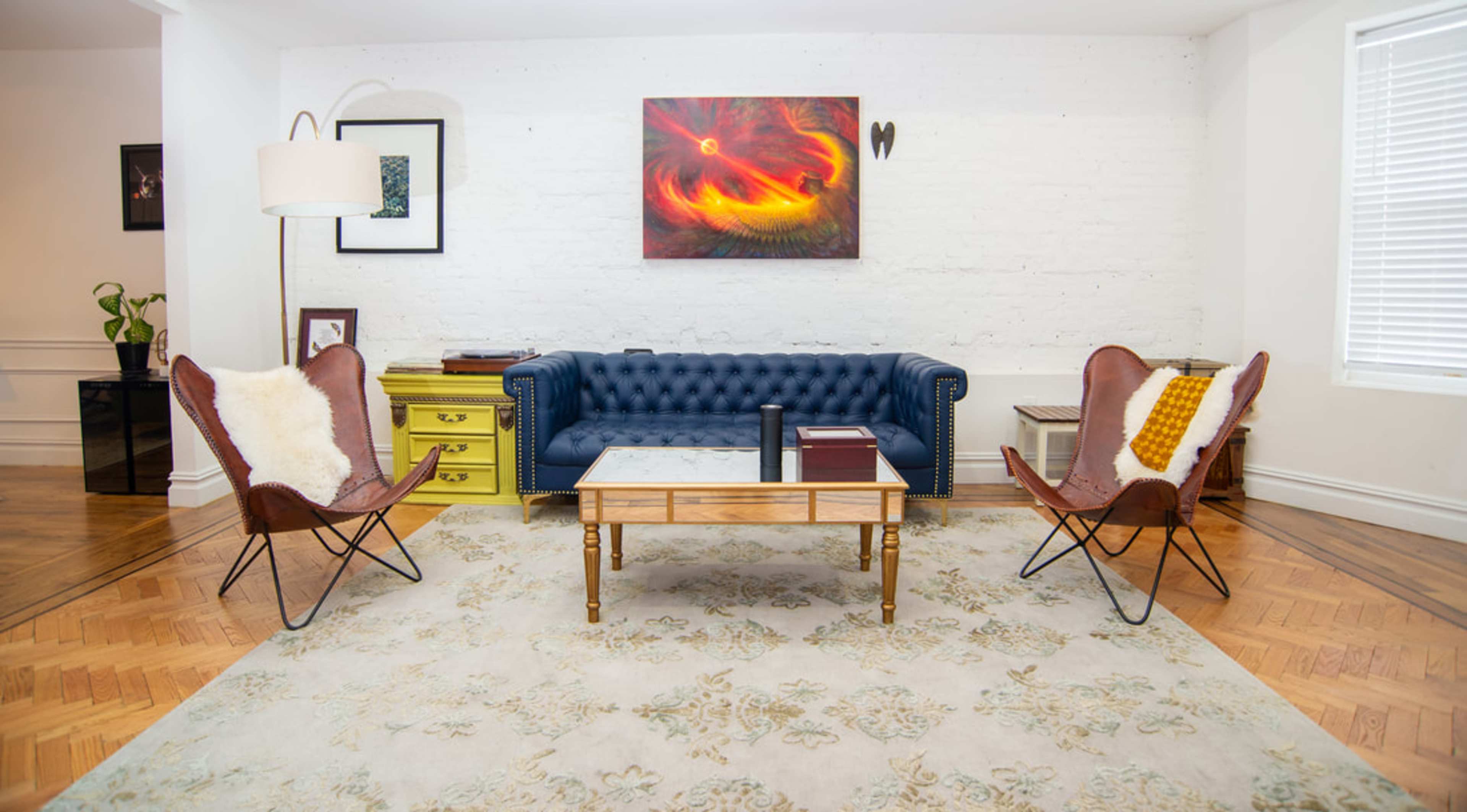 A stylish living room featuring a blue tufted sofa, two brown butterfly chairs, a wooden coffee table, and a colorful abstract artwork on the wall.