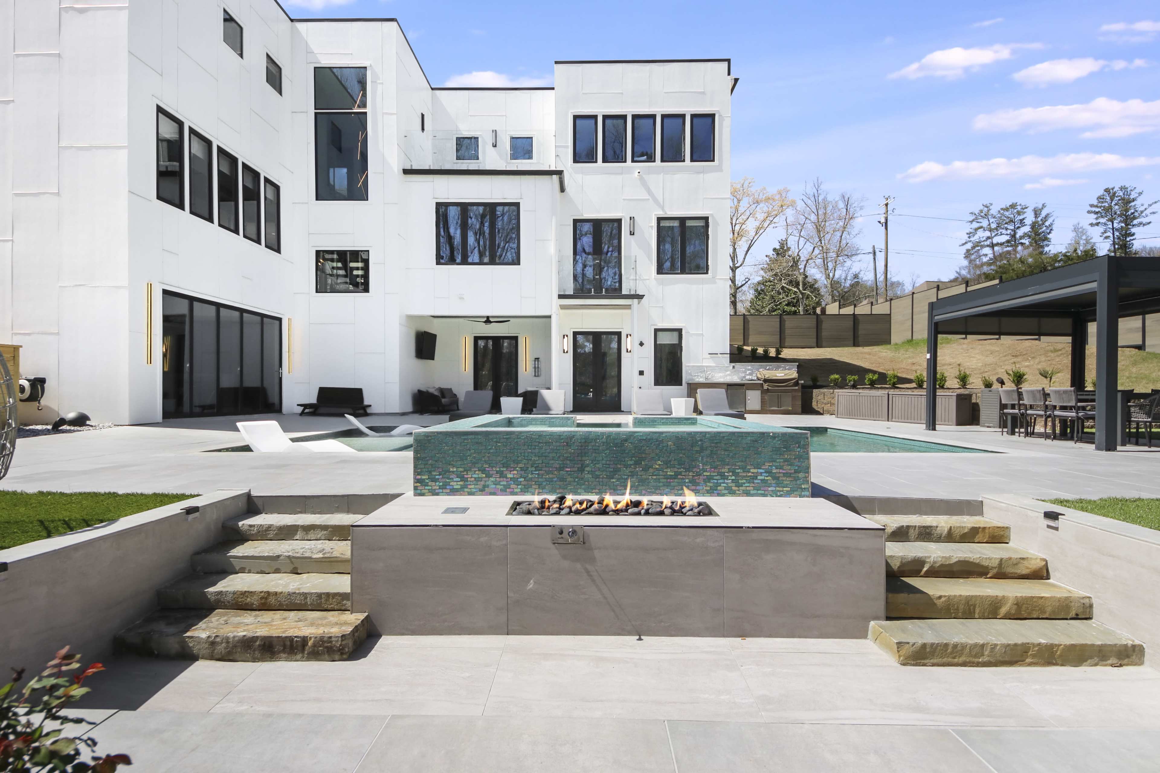 A modern outdoor patio area featuring a swimming pool, a fire pit with a stone surround, and a contemporary house in the background.