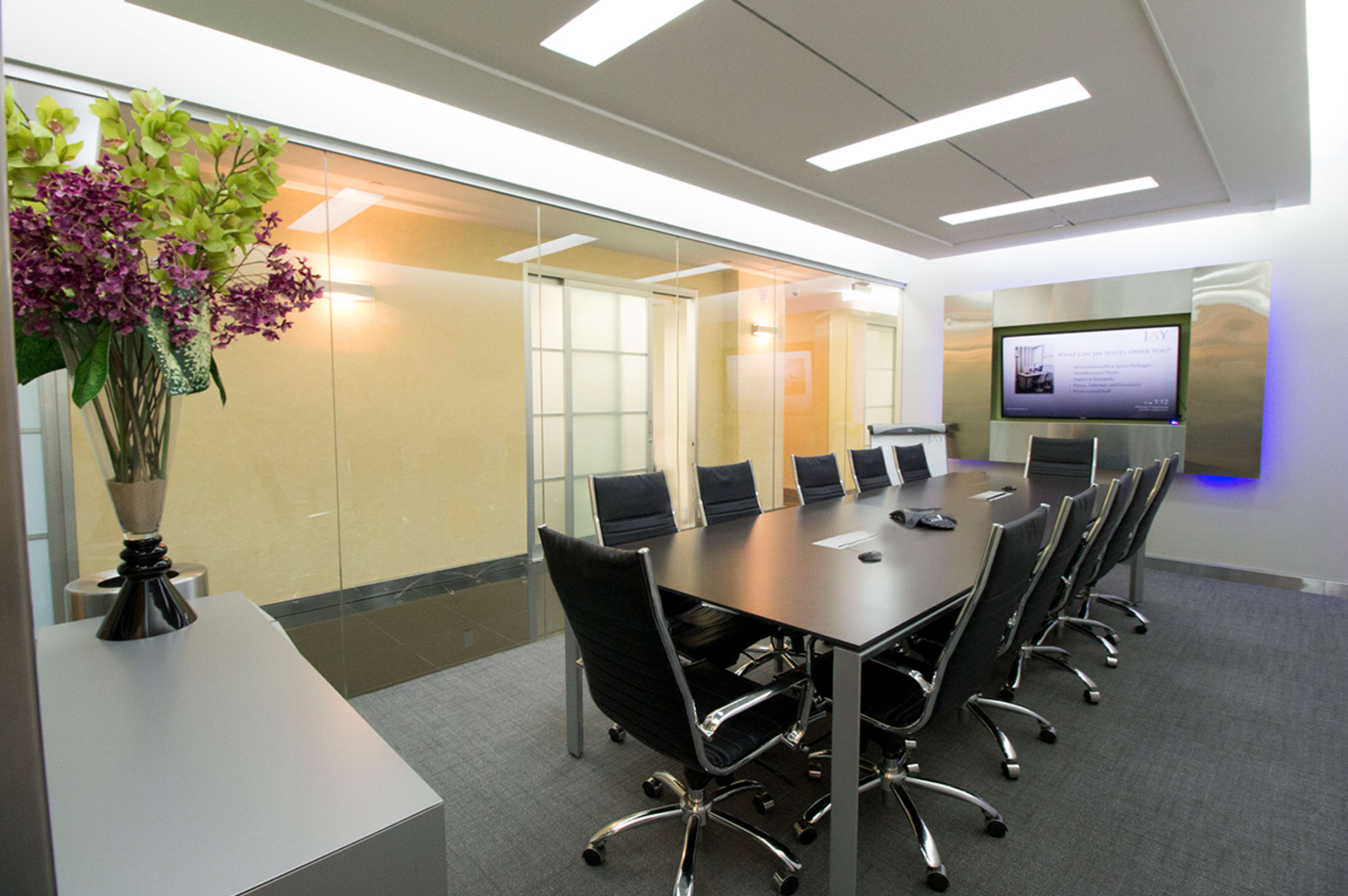 A modern conference room features a large table surrounded by black leather chairs, glass walls, and a wall-mounted screen.