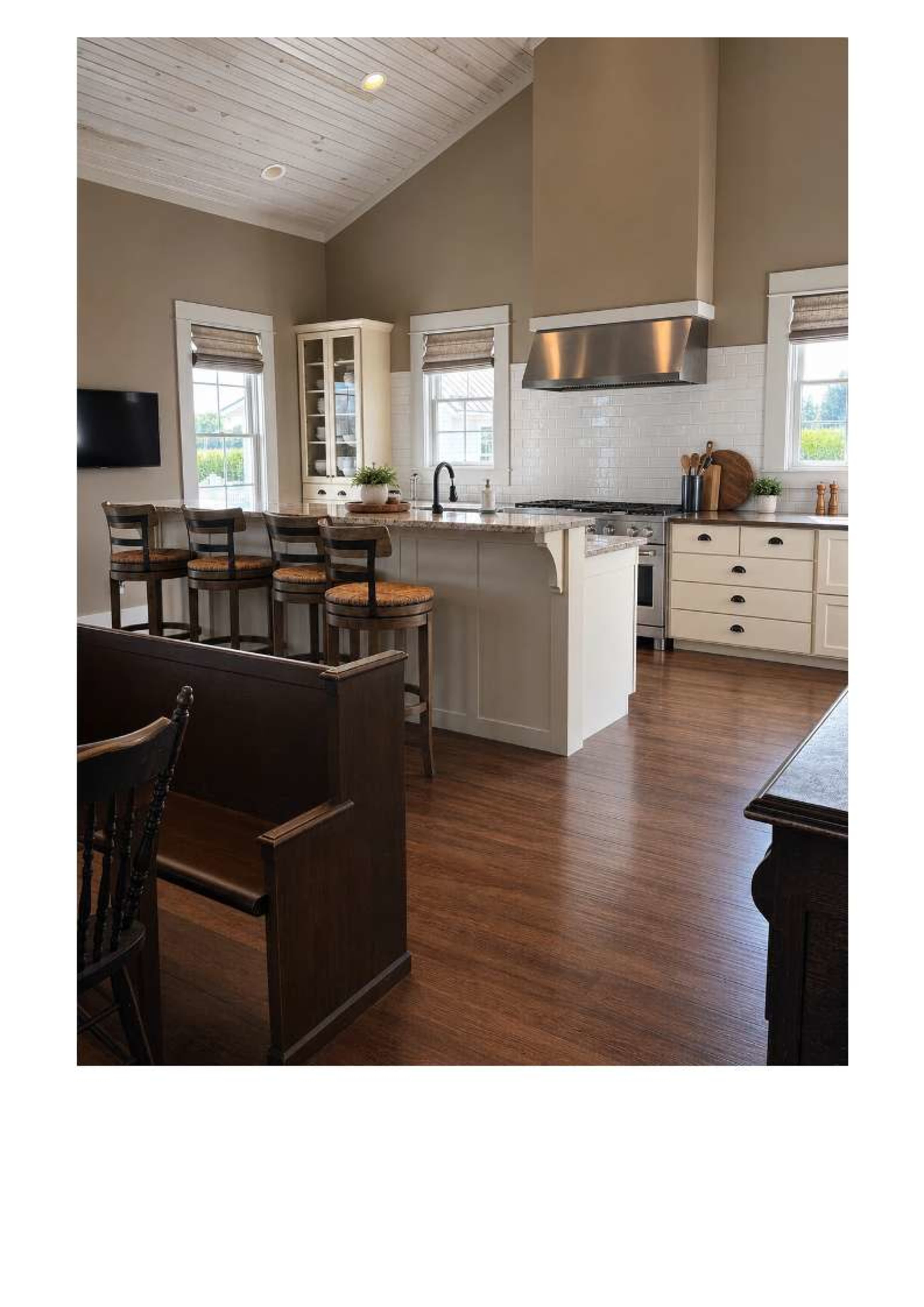 The image shows a modern kitchen with a large island, bar stools, and a combination of light and dark cabinetry, featuring a stainless steel range hood and a large window.