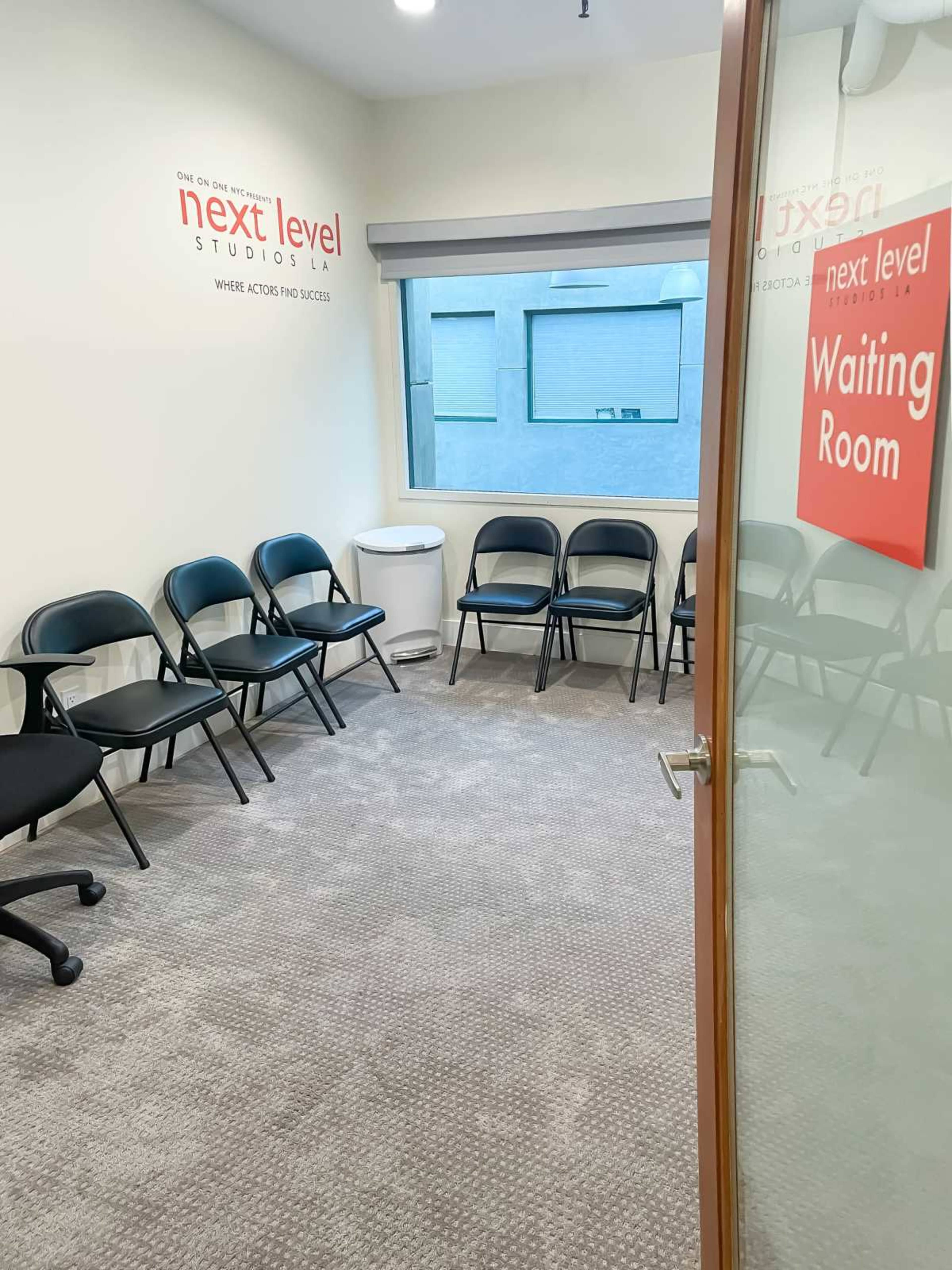 The image shows a waiting room with several black chairs arranged around a carpeted floor, featuring a sign for "Next Level Studios LA" on the wall.