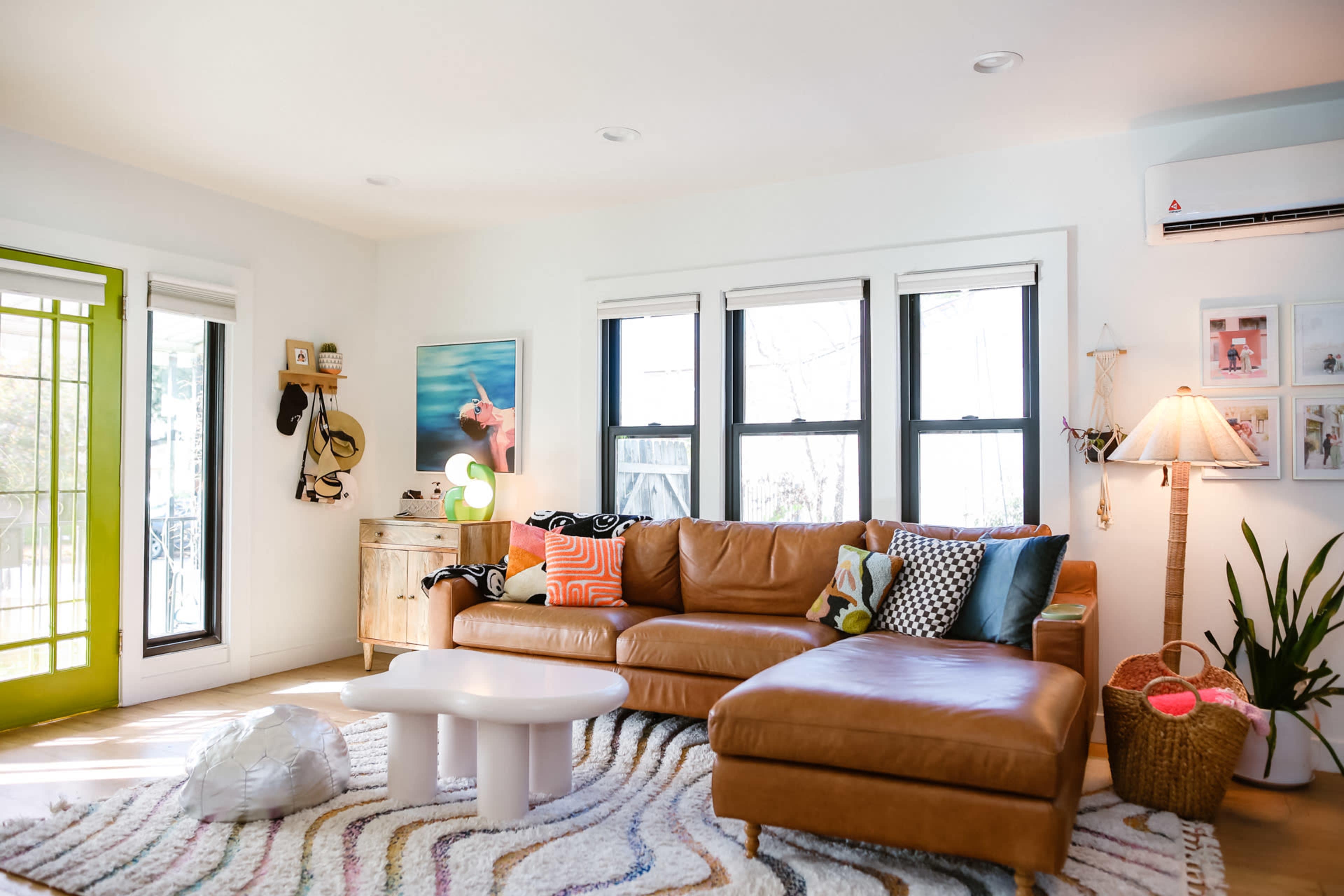 The living room features a brown leather sectional sofa, a white coffee table, and vibrant decor, with large windows allowing natural light to enter.