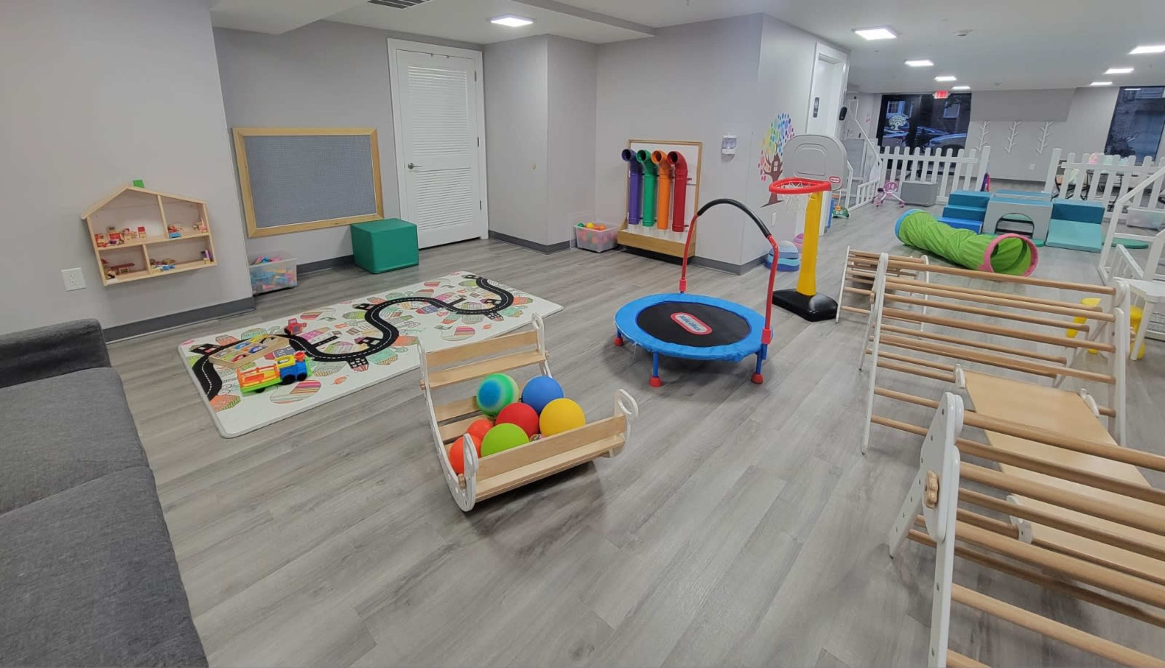 A brightly lit indoor play area filled with various toys, including a train set, colorful balls, and a small trampoline, with soft play equipment scattered around.
