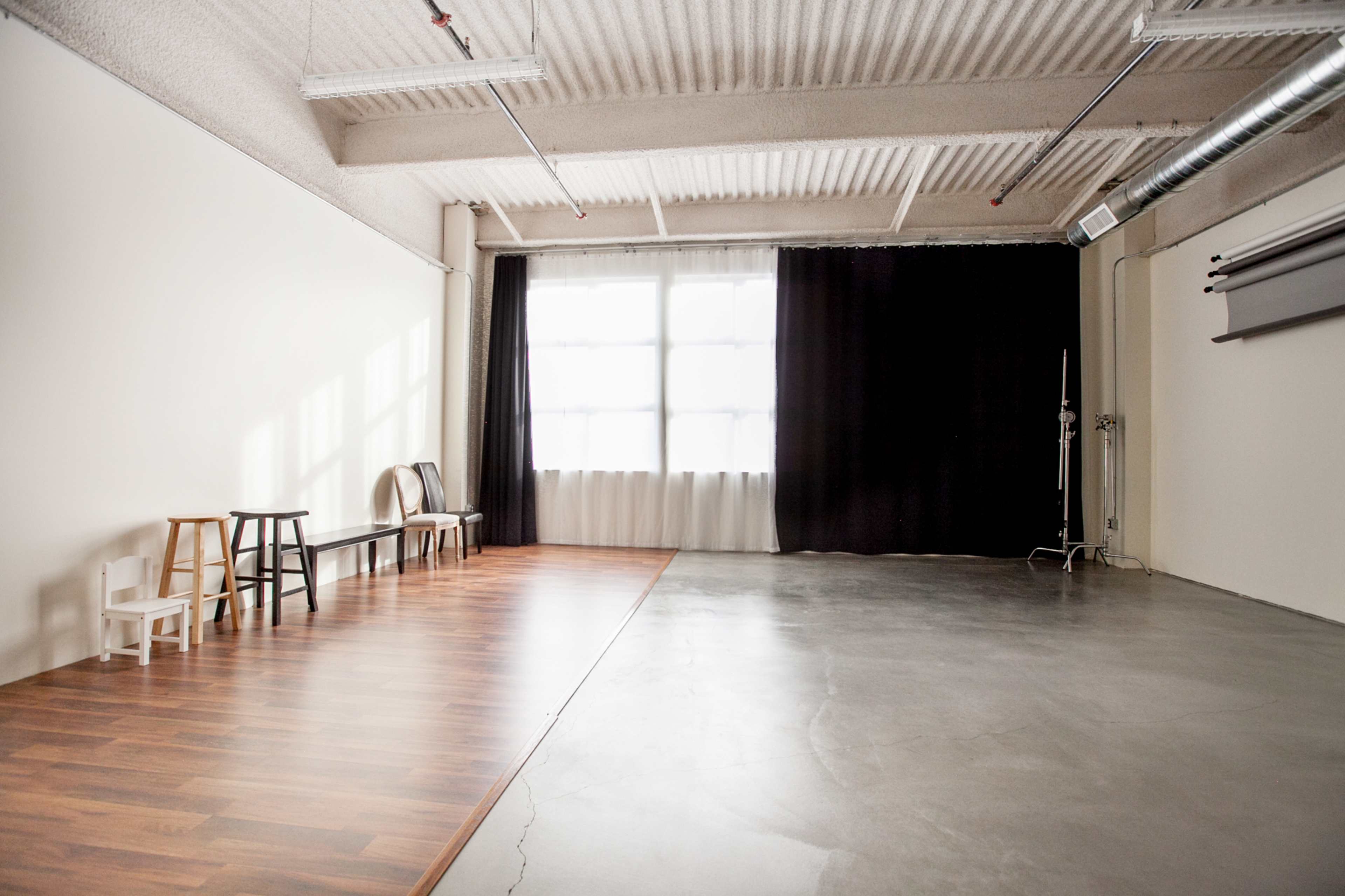 An empty studio space with a wooden floor on one side and a concrete floor on the other, featuring a large window and black curtains.