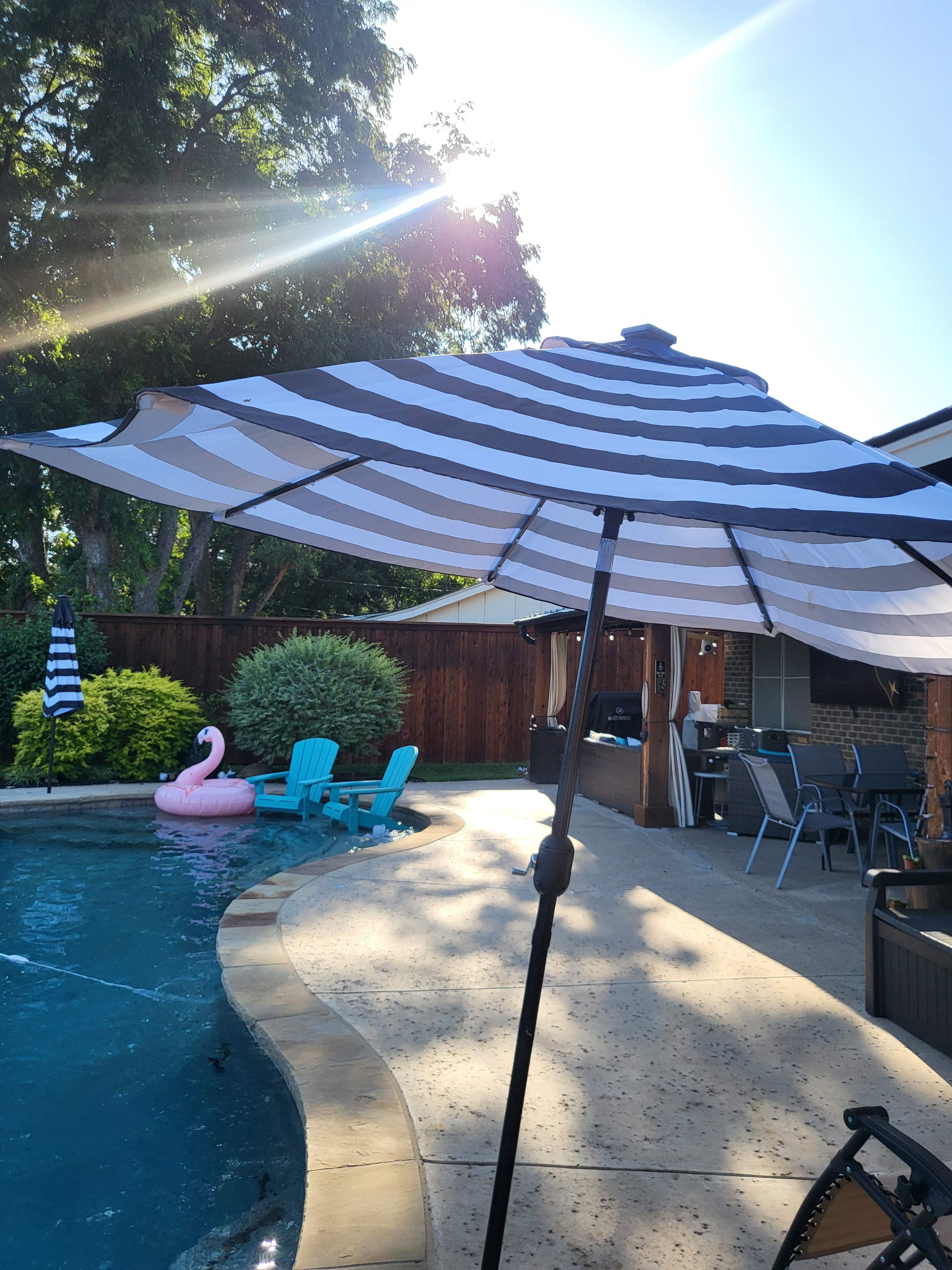 A striped umbrella shades a poolside area featuring a pink flamingo float and blue patio chairs under bright sunlight.