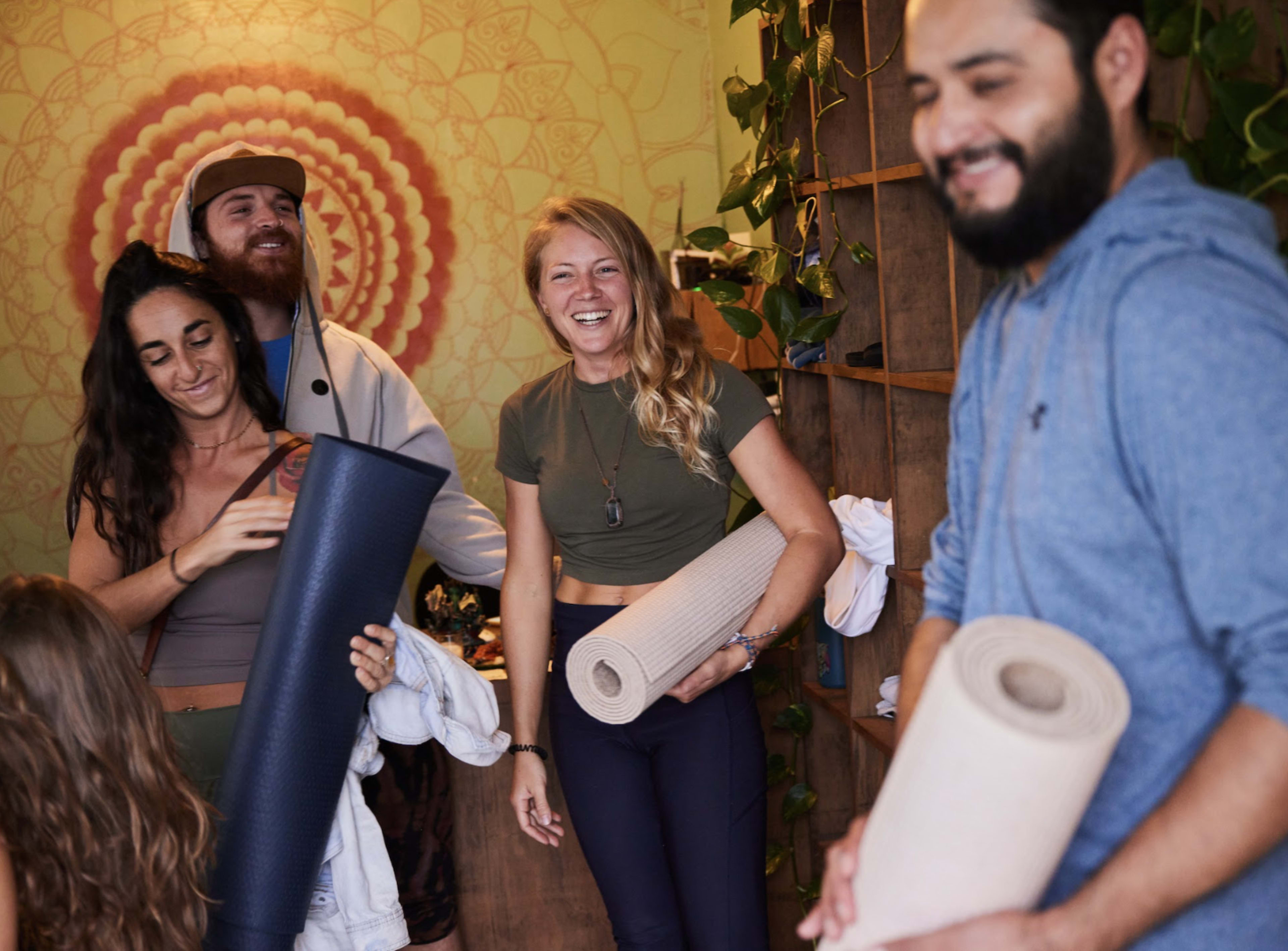 A group of five people stands in a room with plants, each holding a yoga mat and smiling at one another.
