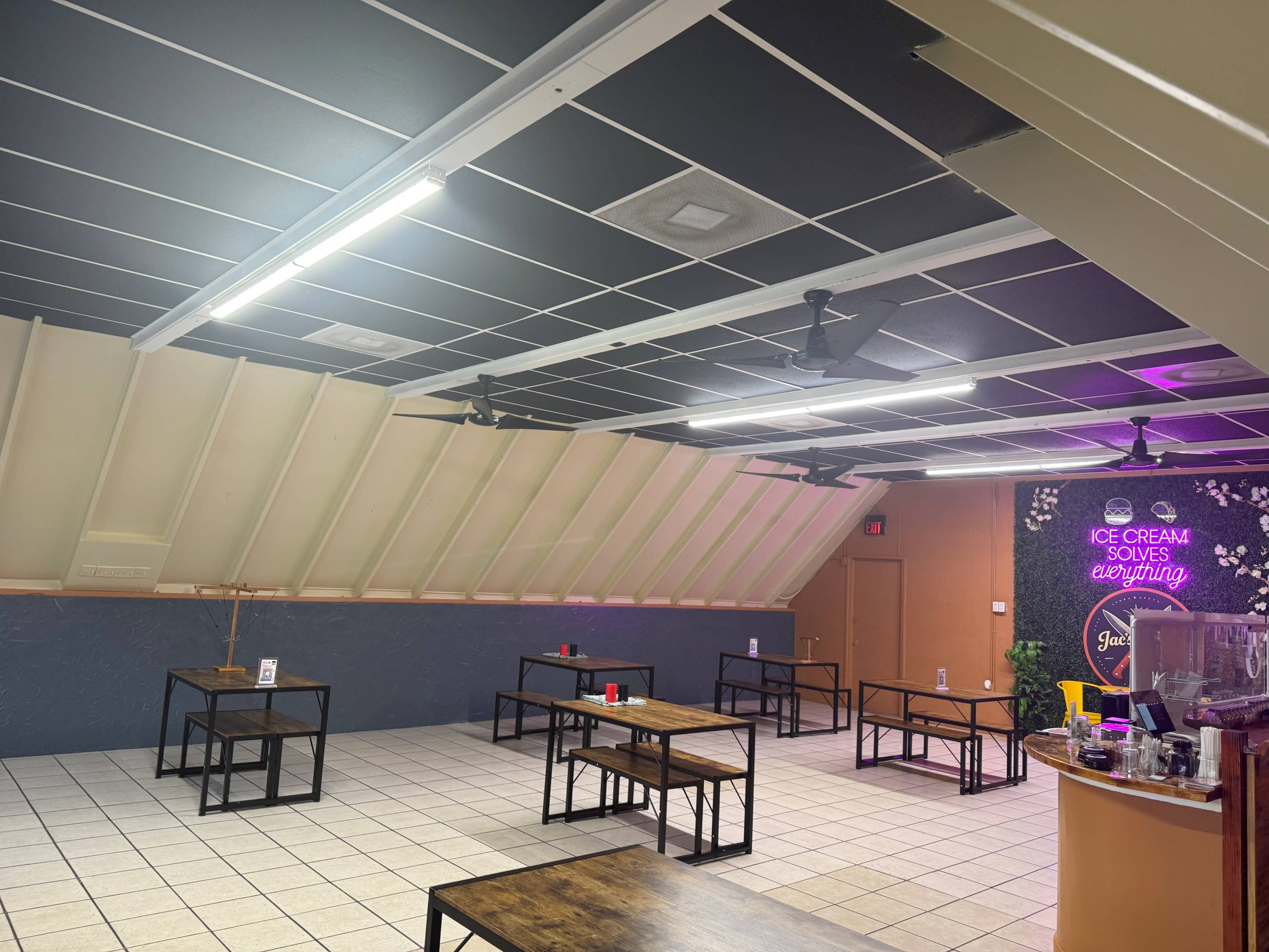The image shows an indoor space with a sloped ceiling, featuring several wooden tables and a counter area on one side, and a neon sign that reads "ICE CREAM SOLVES EVERYTHING."