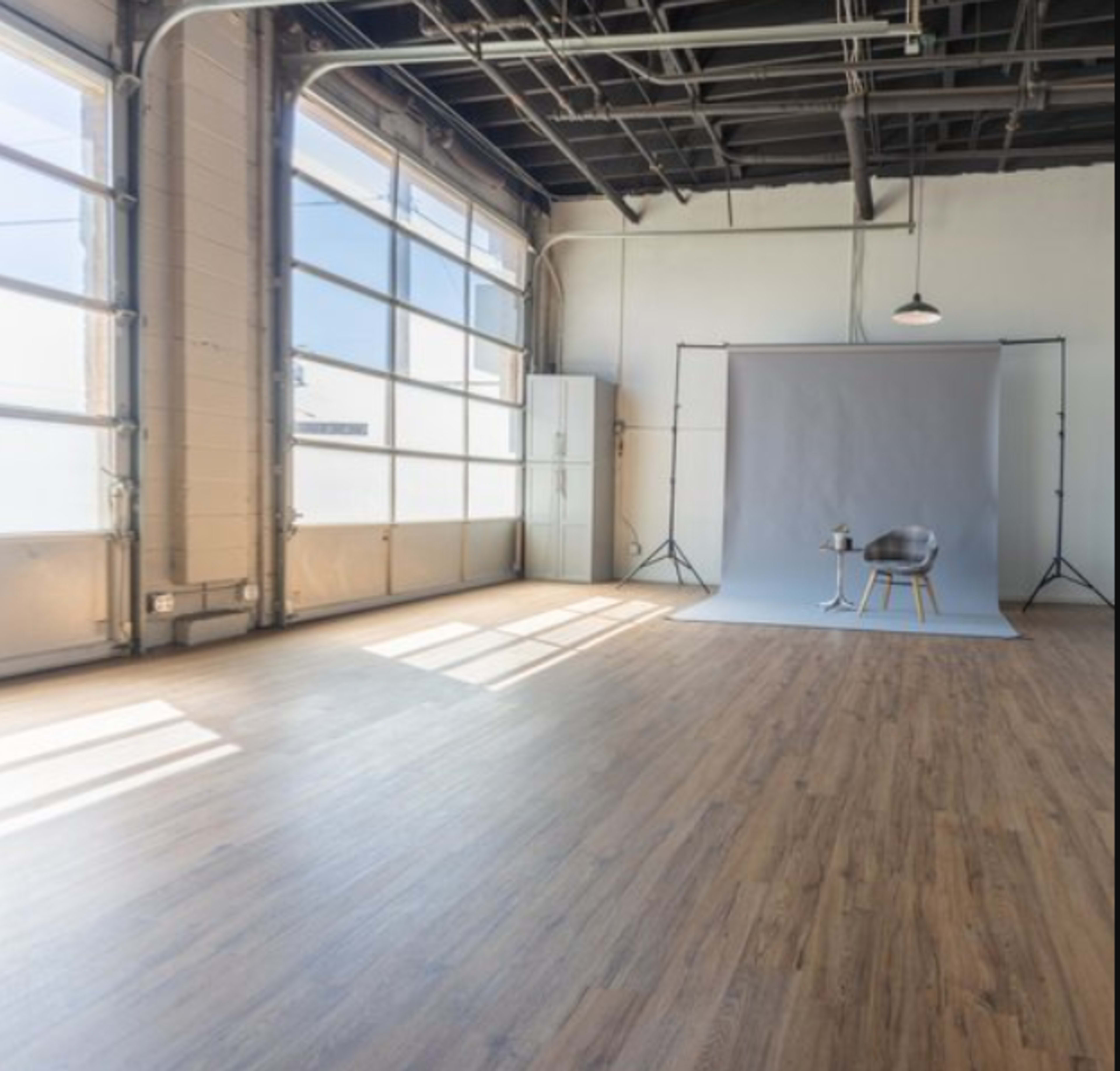 The image shows a spacious, well-lit studio with large windows, wooden flooring, and a simple backdrop setup featuring a chair and a small table.