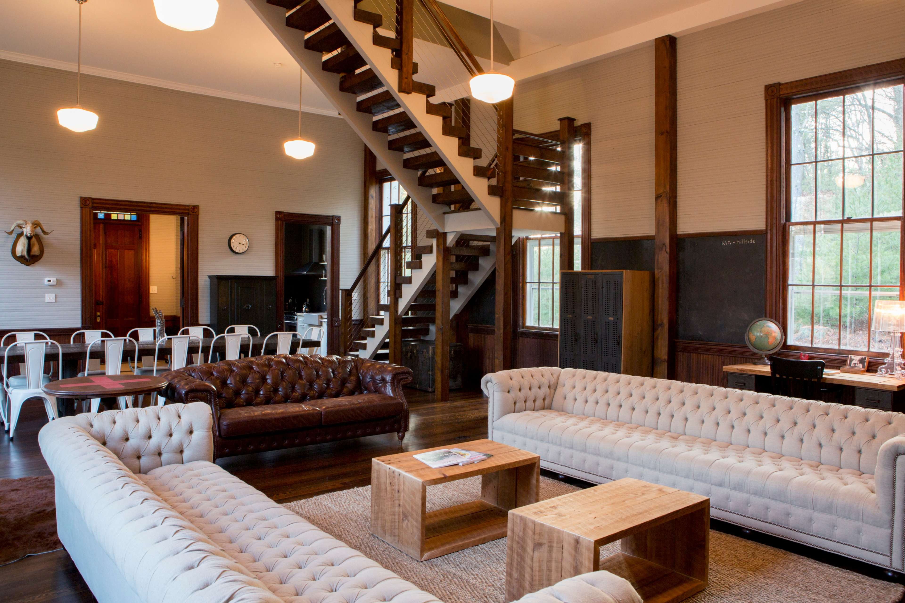 The room features a spacious interior with two tufted sofas, wooden furniture, and a staircase leading to an upper level.