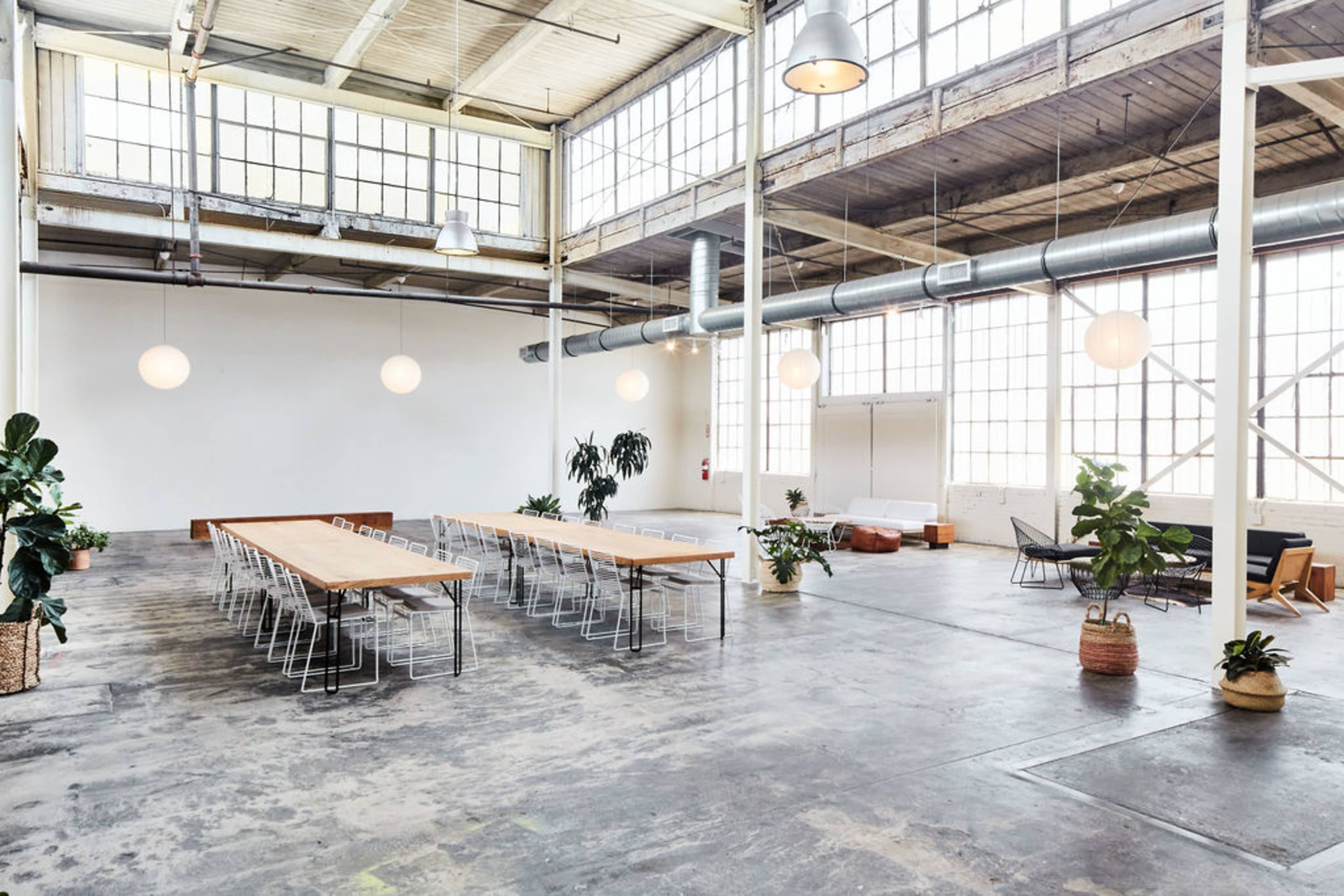The image shows a spacious, industrial-style room with large windows, a long wooden table surrounded by metal chairs, and various plants positioned around the area.