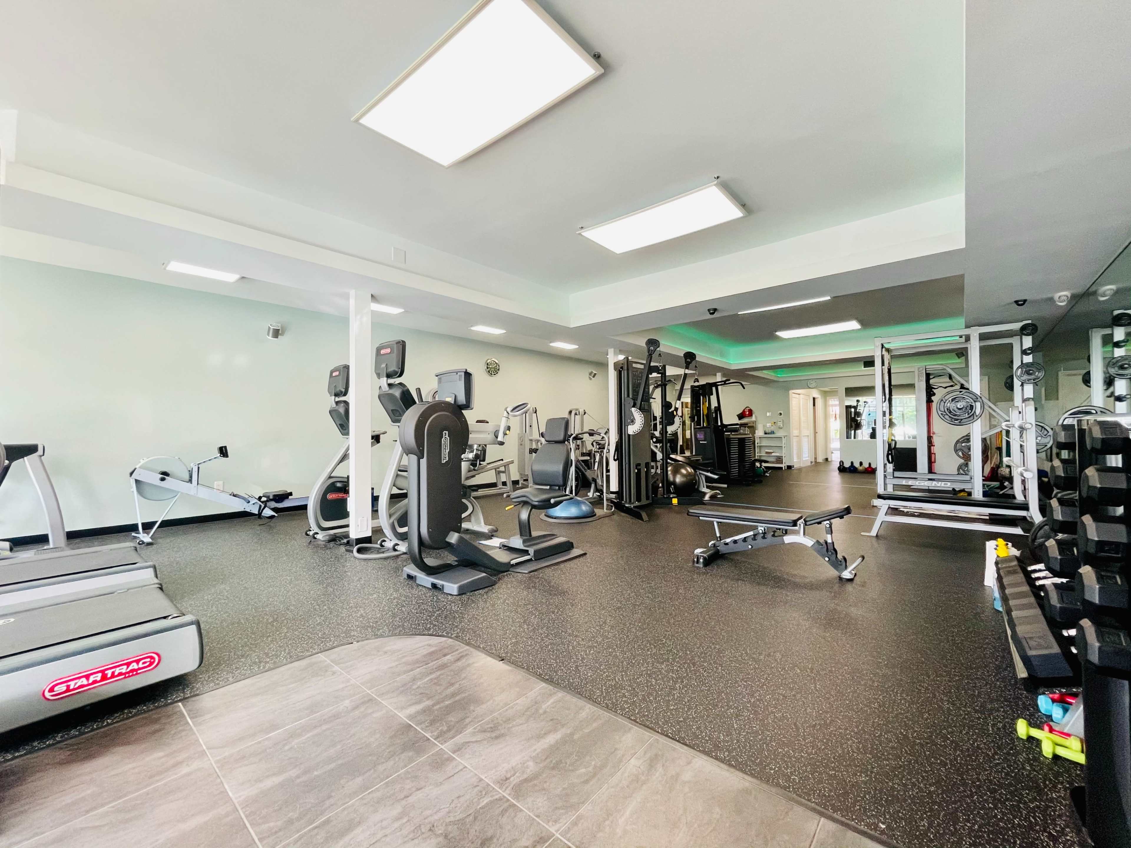 The image shows a fitness gym with various exercise machines, free weights, and a treadmill arranged on a rubberized floor.