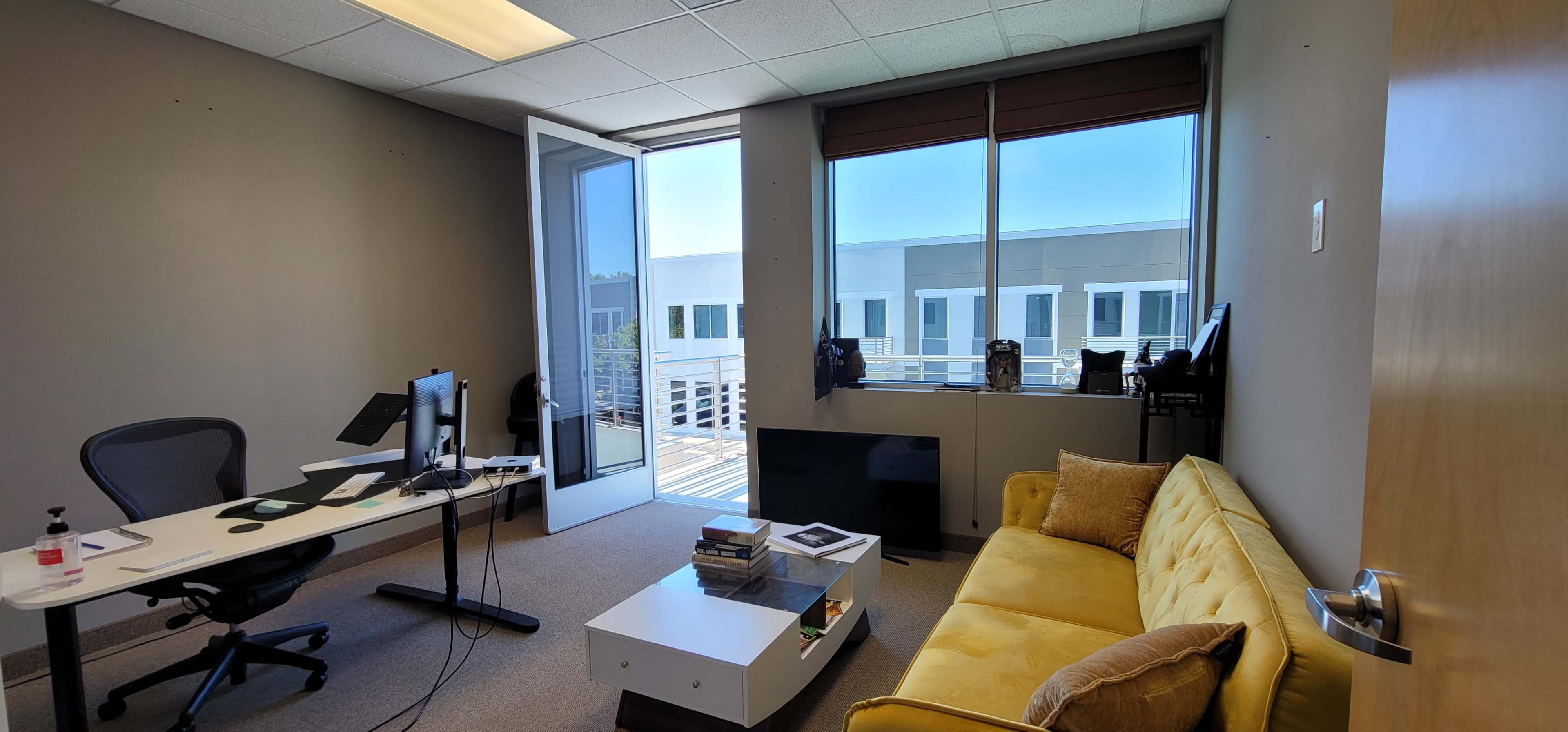 An office space features a yellow couch, a desk with two monitors, and large windows opening to a balcony.