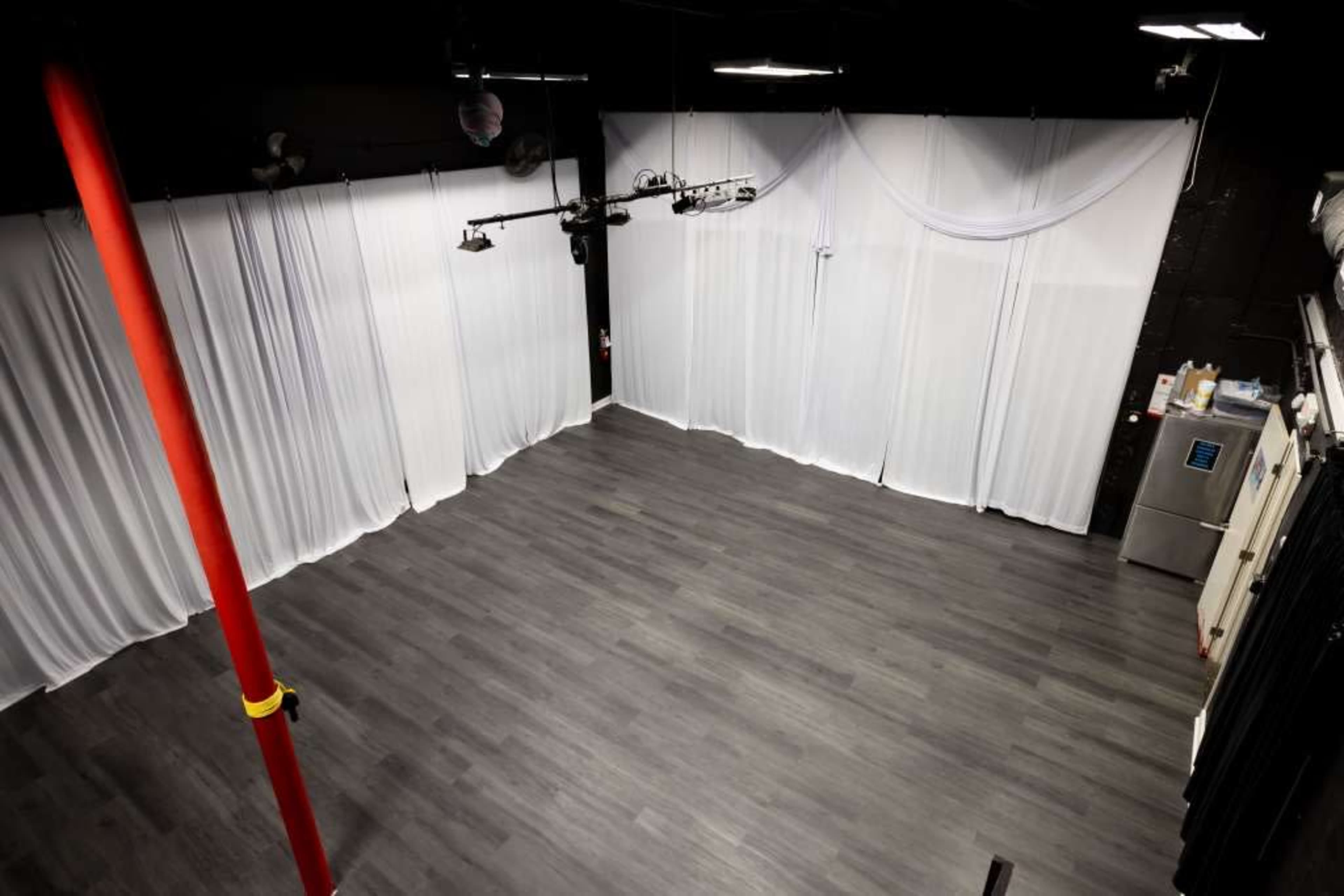 The image shows a spacious, empty studio with white drapes, wooden flooring, and overhead lighting.