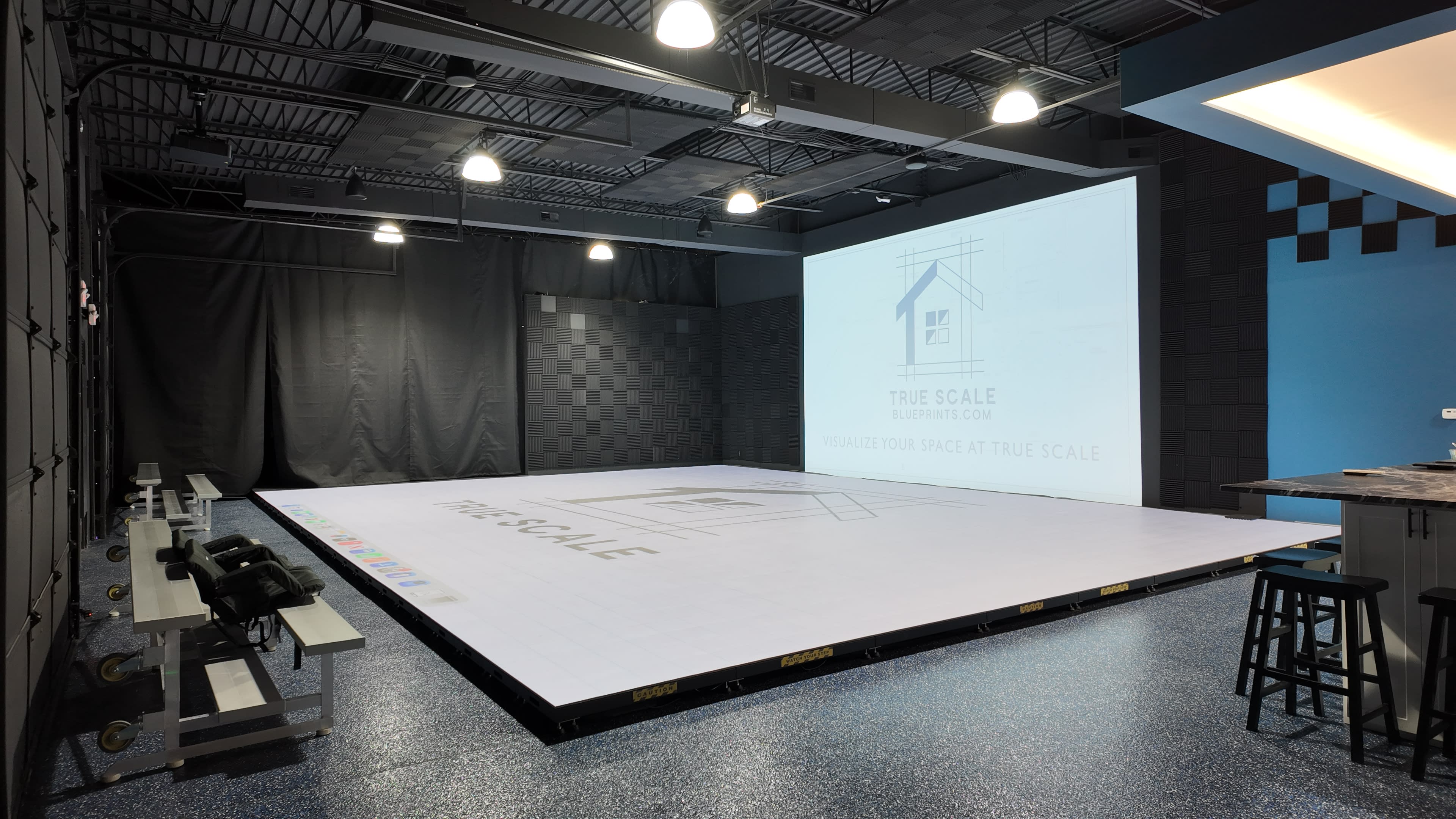 The image shows a spacious room with a large, flat projection surface displaying a design layout, and there are benches and stools along the sides.