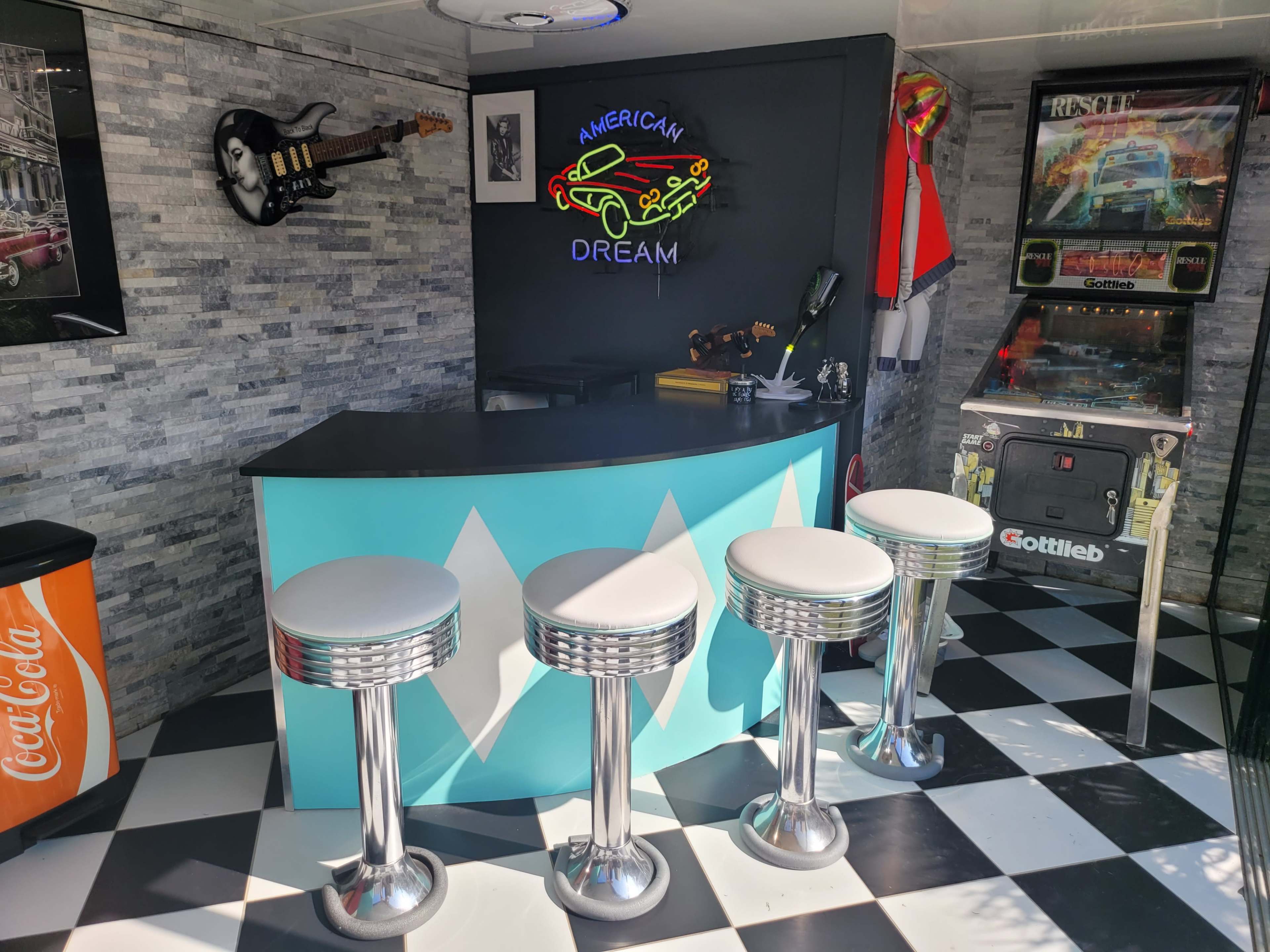 The image shows a retro-themed bar area with a black and white checkered floor, a neon "American Dream" sign, and four chrome bar stools in front of a sleek counter.