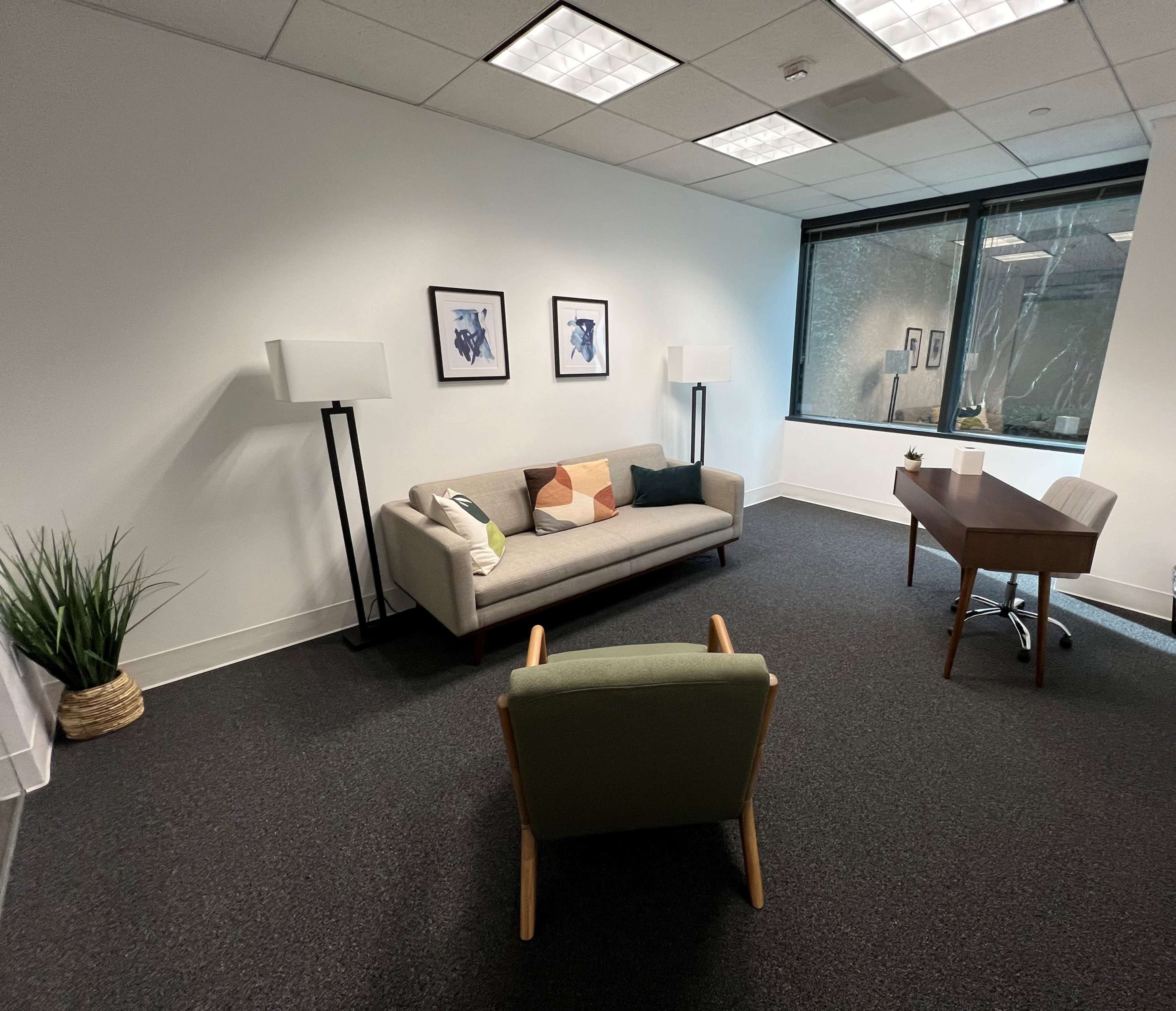 A minimalist office space featuring a gray sofa, a wooden desk with a chair, and two floor lamps against a white wall with two framed pictures.