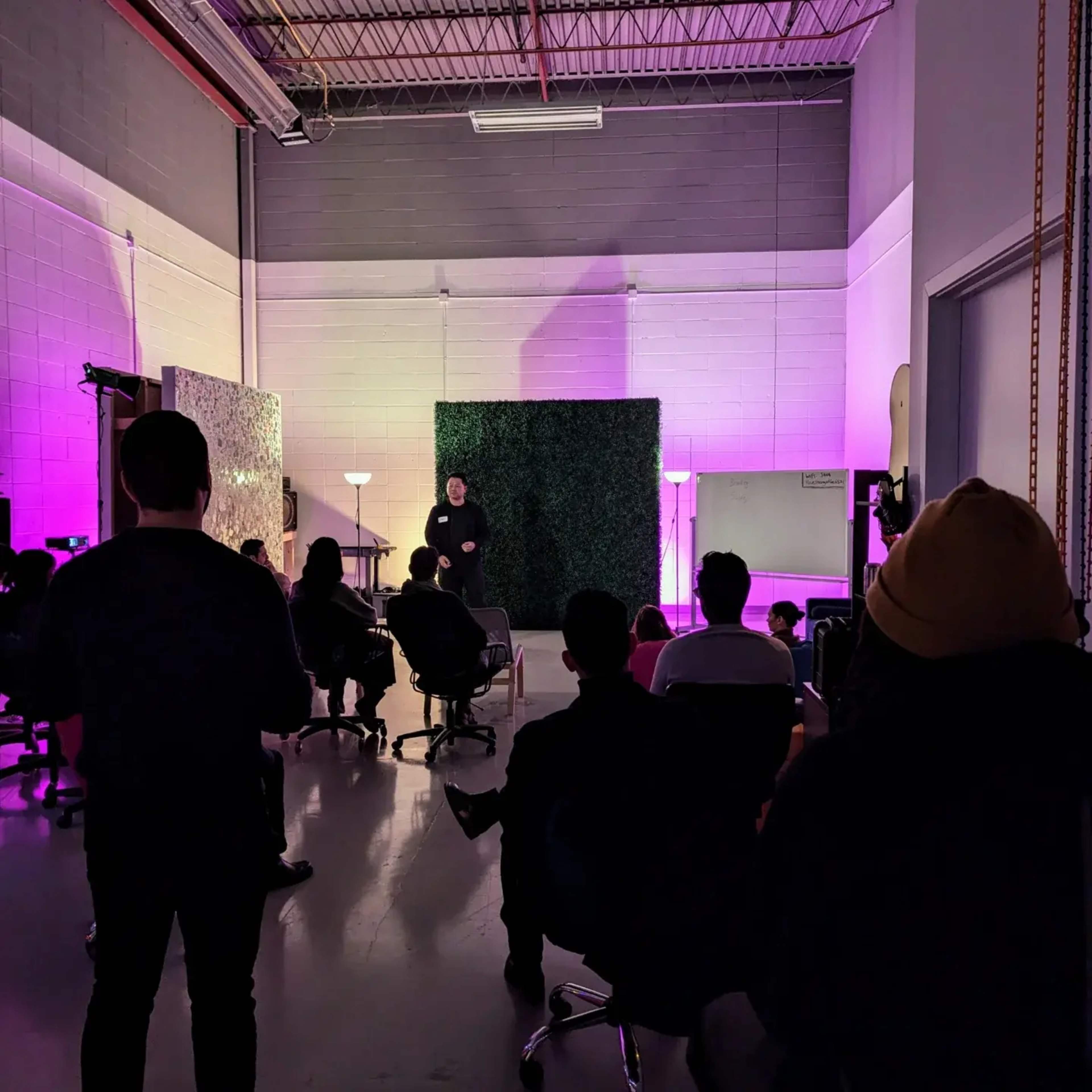 A speaker addresses an audience in a dimly lit room decorated with green plants and purple lighting.