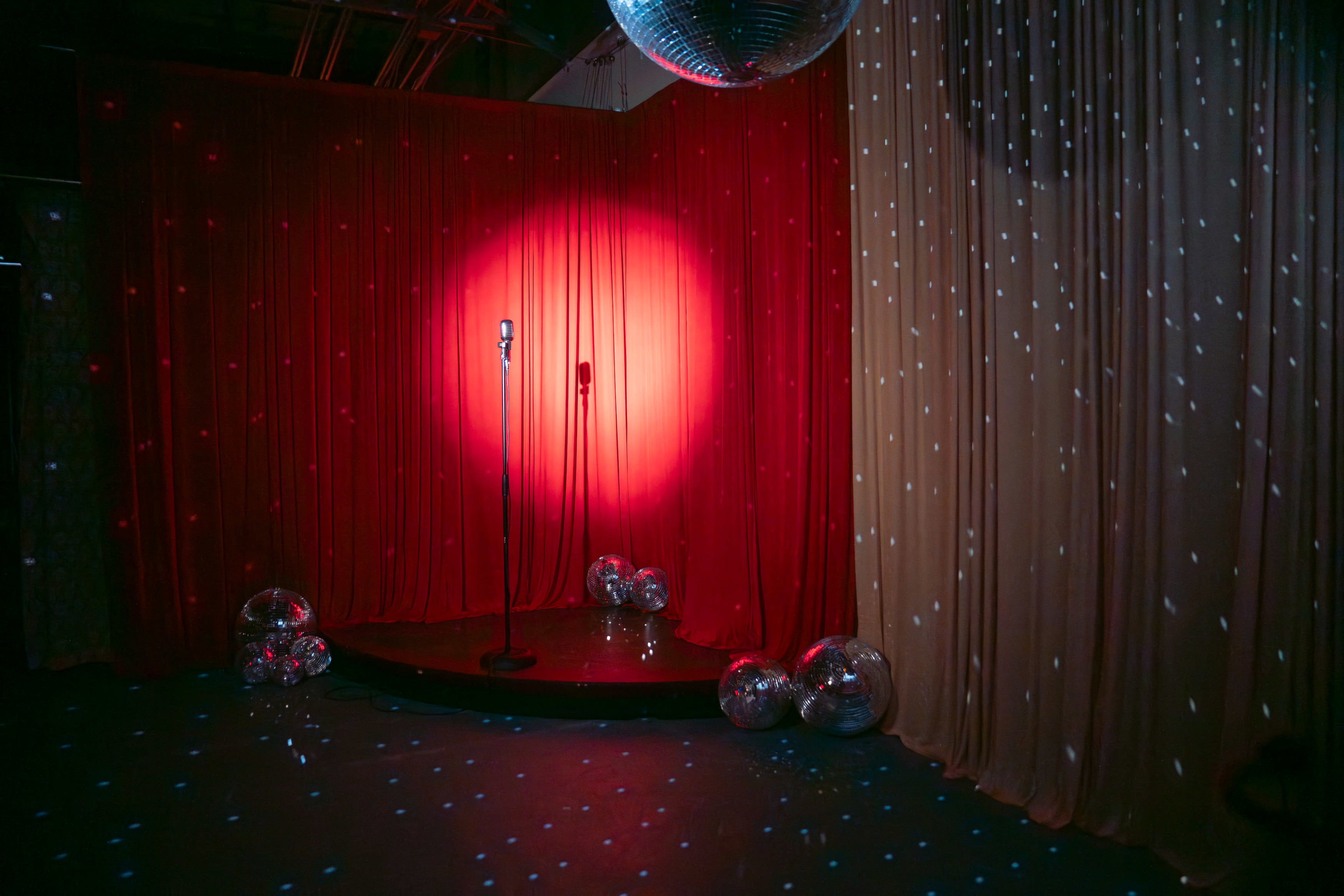 The image shows a stage with a microphone on a circular platform surrounded by shiny disco balls and draped red and cream curtains, illuminated by colorful lights.