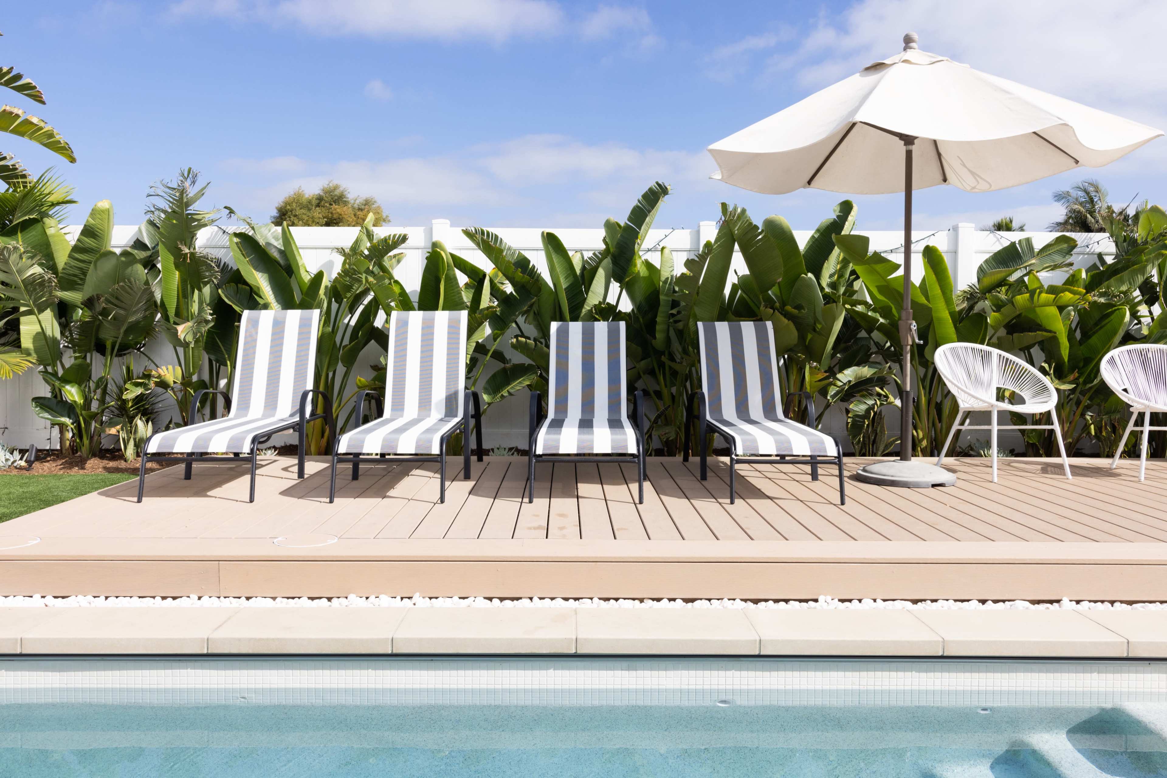 A row of four striped lounge chairs and a white umbrella sits beside a swimming pool, surrounded by tall green plants.