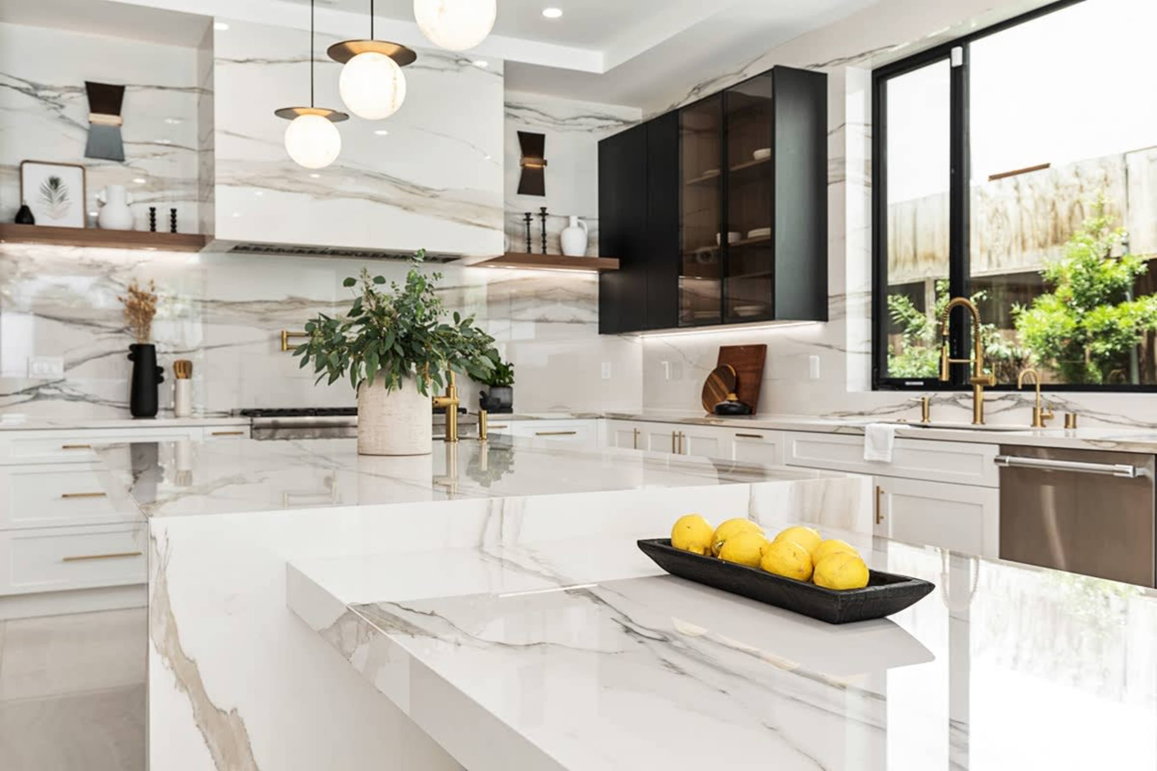A modern kitchen features a spacious marble island with a black tray holding lemons, sleek cabinetry, and large windows allowing natural light.