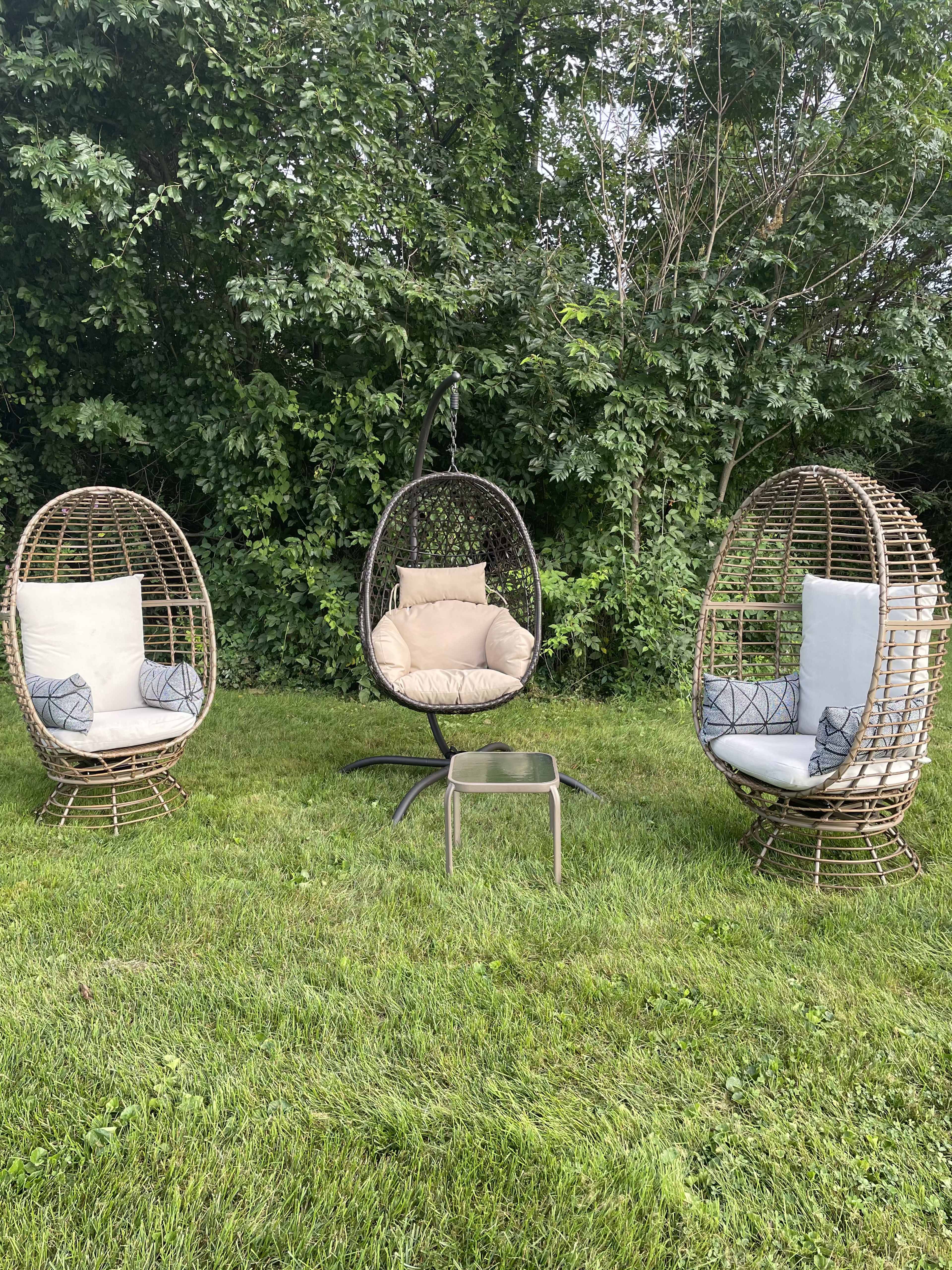 Three woven garden chairs, including a hanging chair, arranged on a grassy area surrounded by trees.