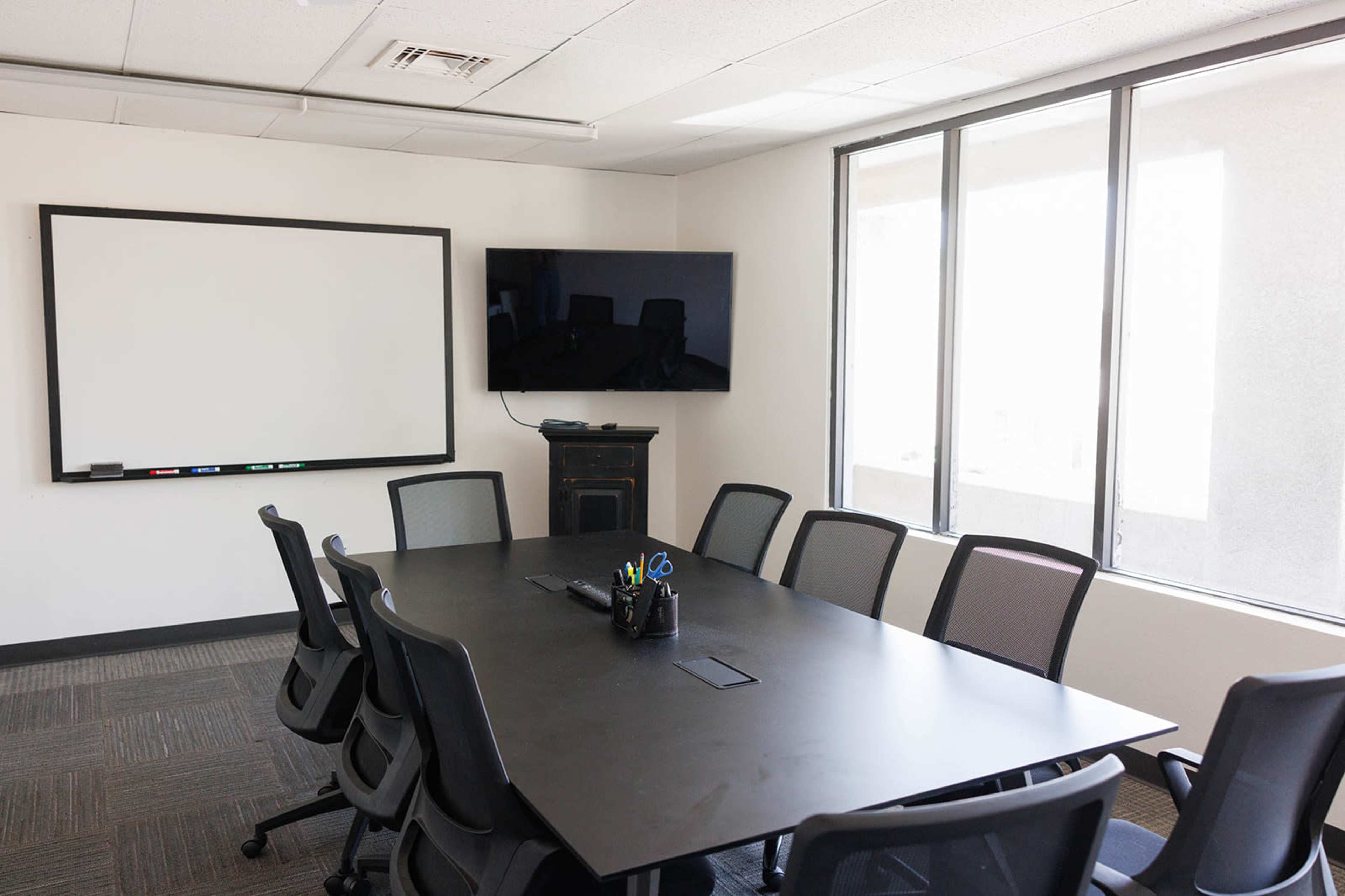 The image shows a modern conference room with a large black table, several ergonomic chairs, a whiteboard, and a wall-mounted television.
