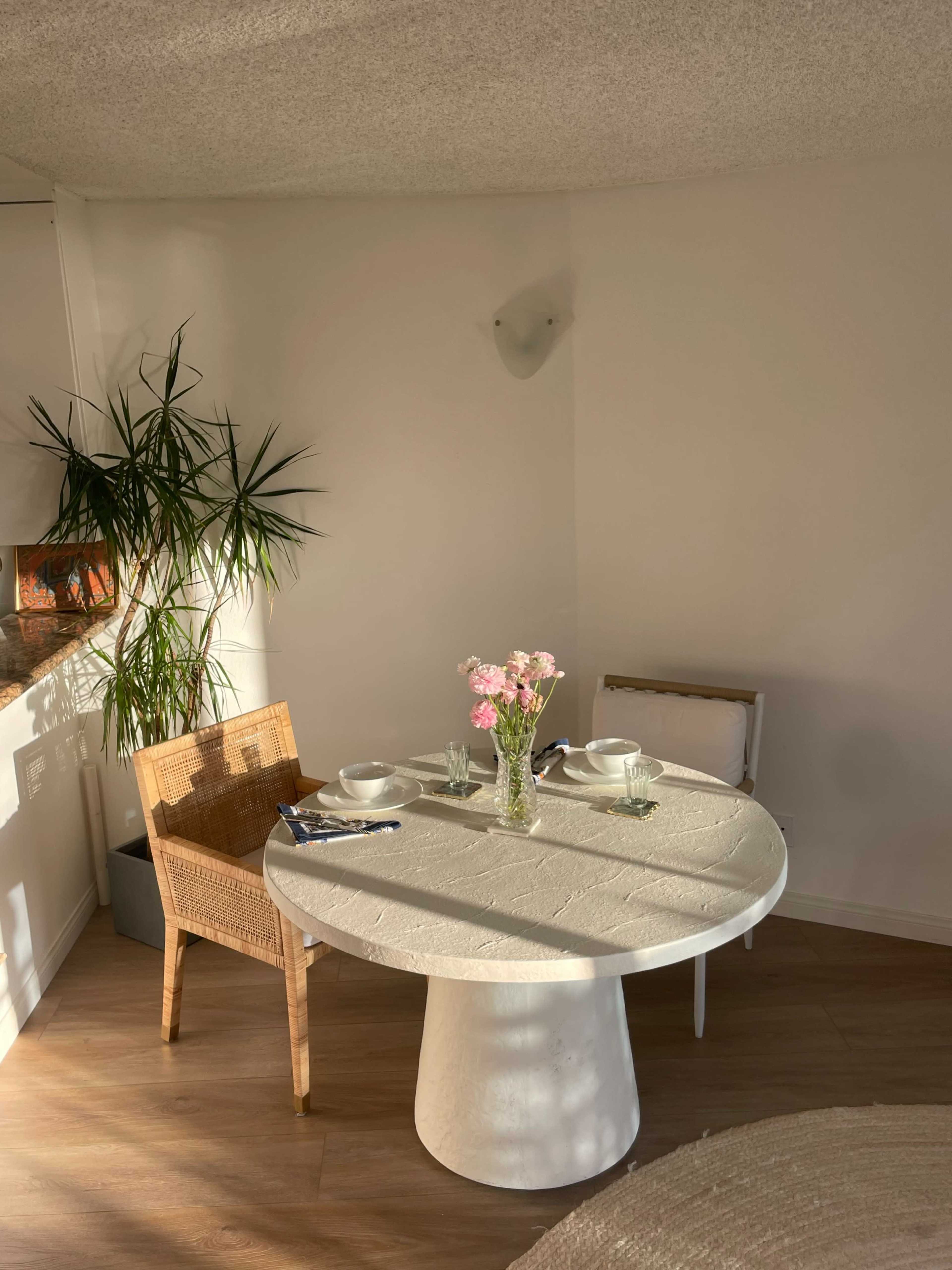 A round white table is set for two with a vase of flowers in the center, accompanied by a potted plant and a single chair beside it in a well-lit dining area.