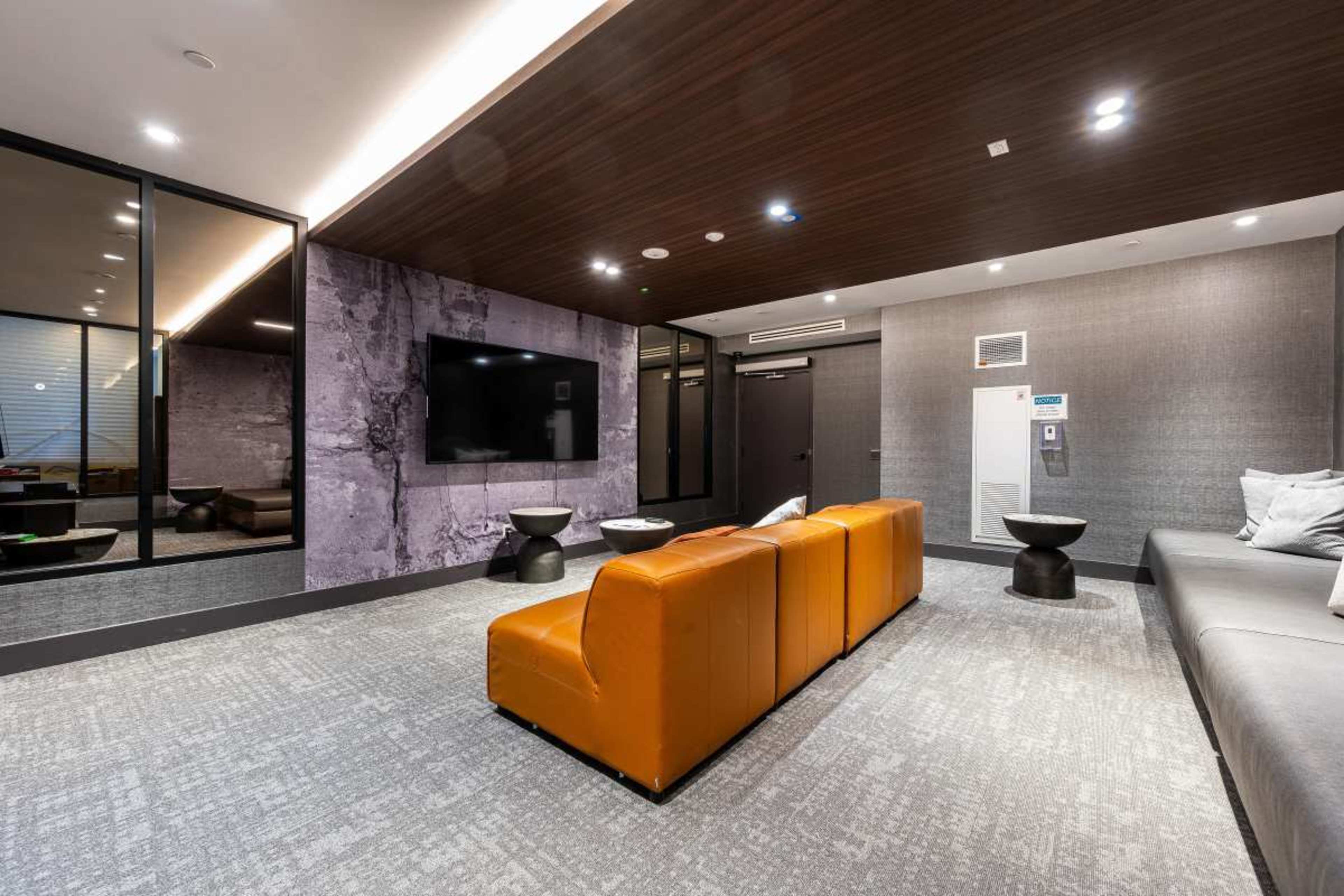 The image shows a modern lounge area with a mix of orange and gray seating, a large wall-mounted television, and a sleek design featuring glass and textured walls.