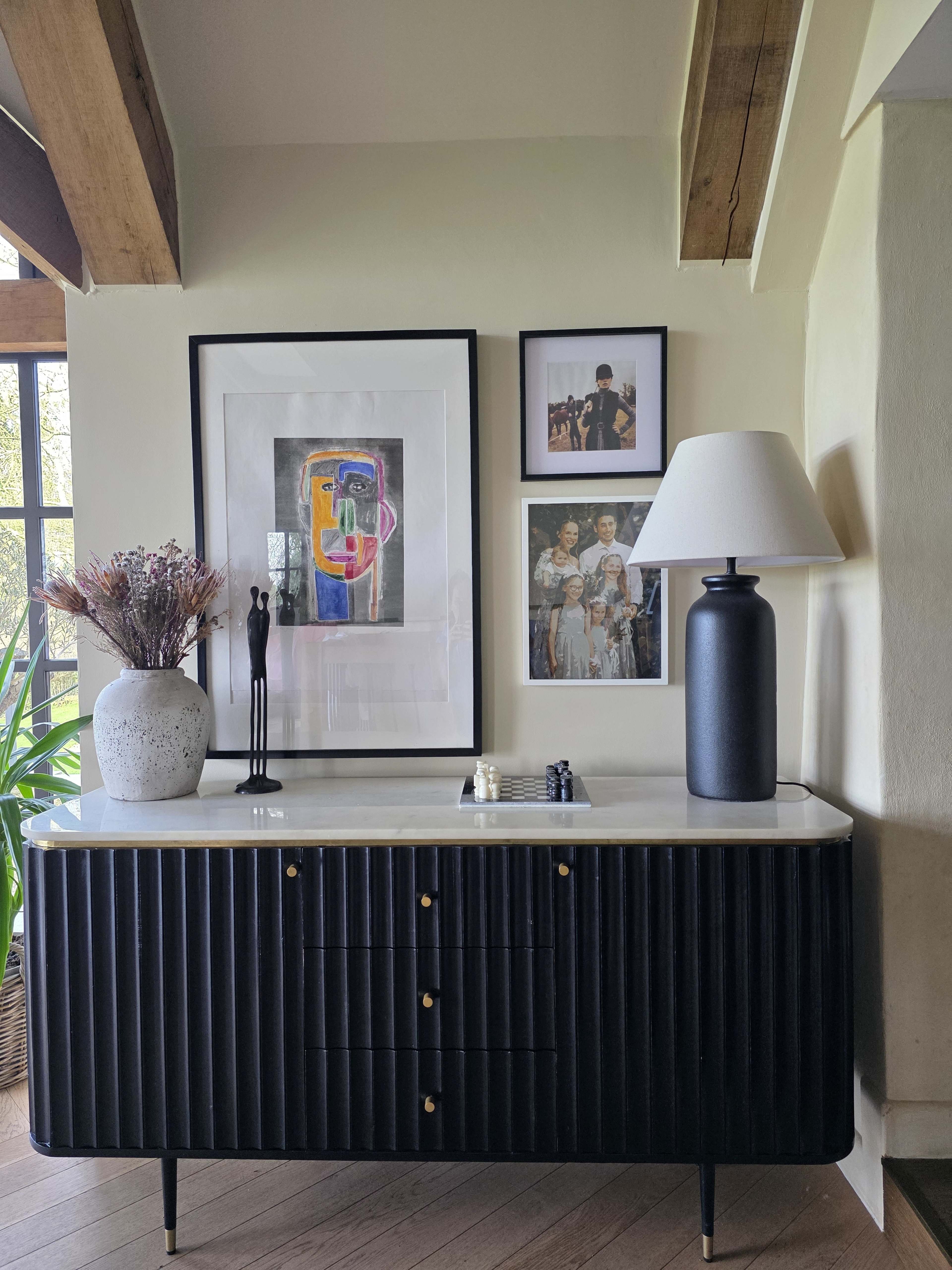 A modern, black sideboard with a marble top is positioned against a light-colored wall, adorned with framed artwork and photographs, alongside a tall black lamp and a decorative vase of flowers.