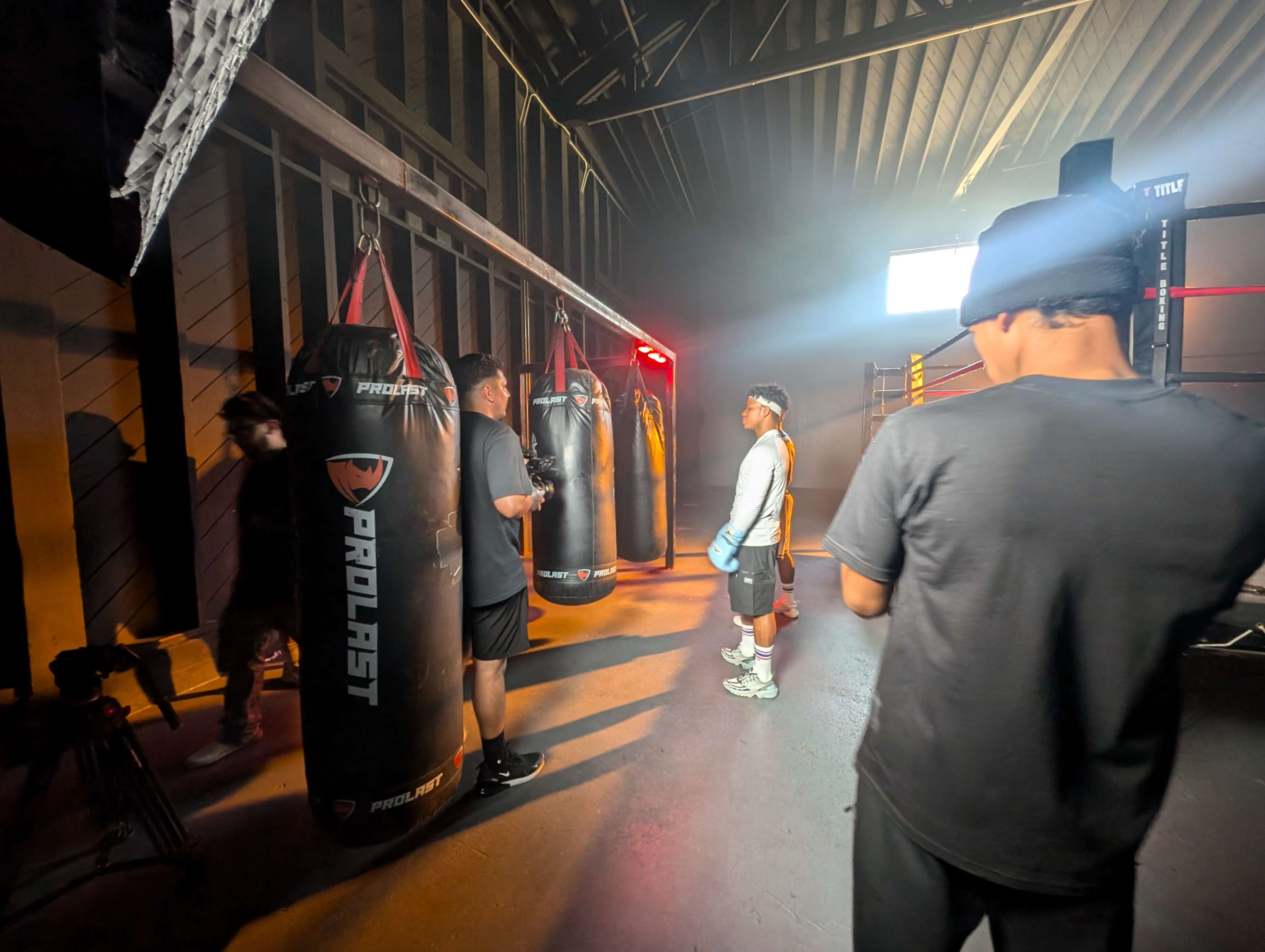 A group of individuals stands in a dimly lit gym with several hanging punching bags, preparing for a boxing training session.