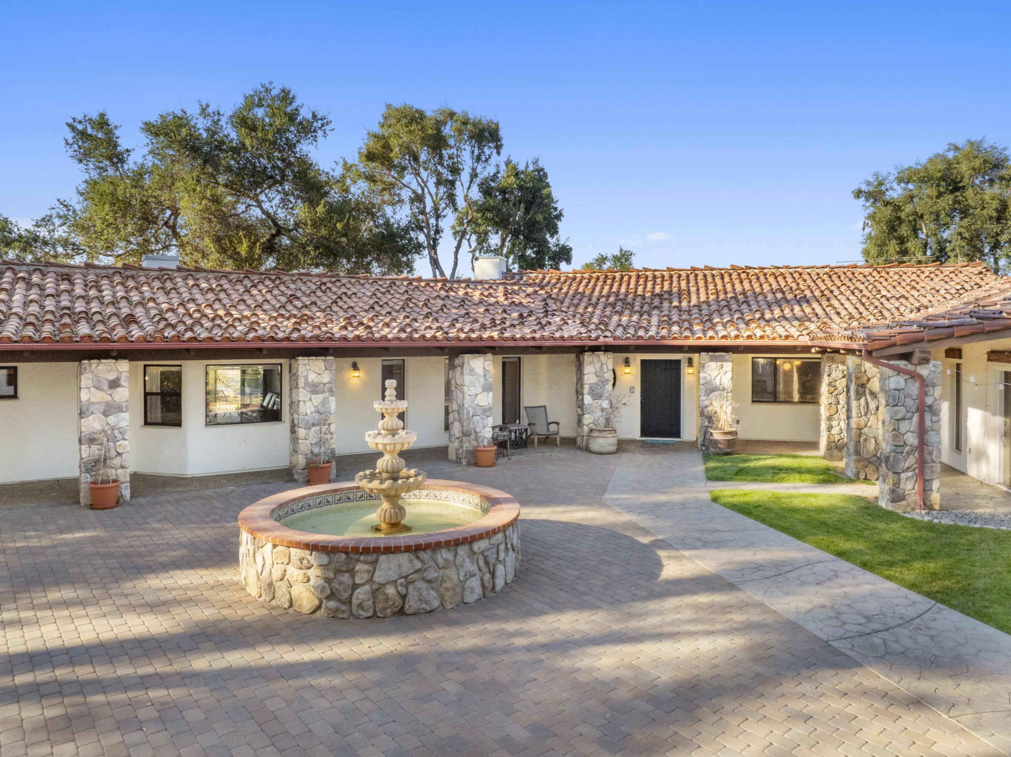 The image shows a rustic courtyard featuring a stone fountain surrounded by a single-story house with a red-tiled roof and paver stone paths.
