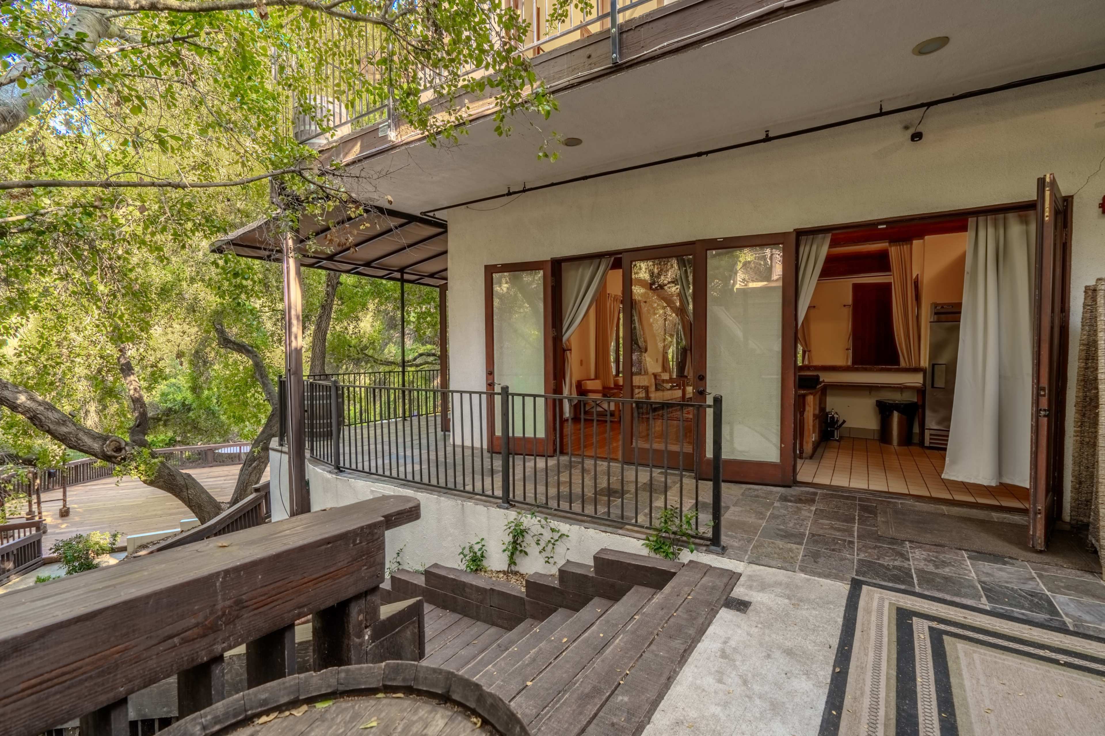 The image shows a spacious patio area leading to a modern indoor space with large glass doors and a surrounding garden.
