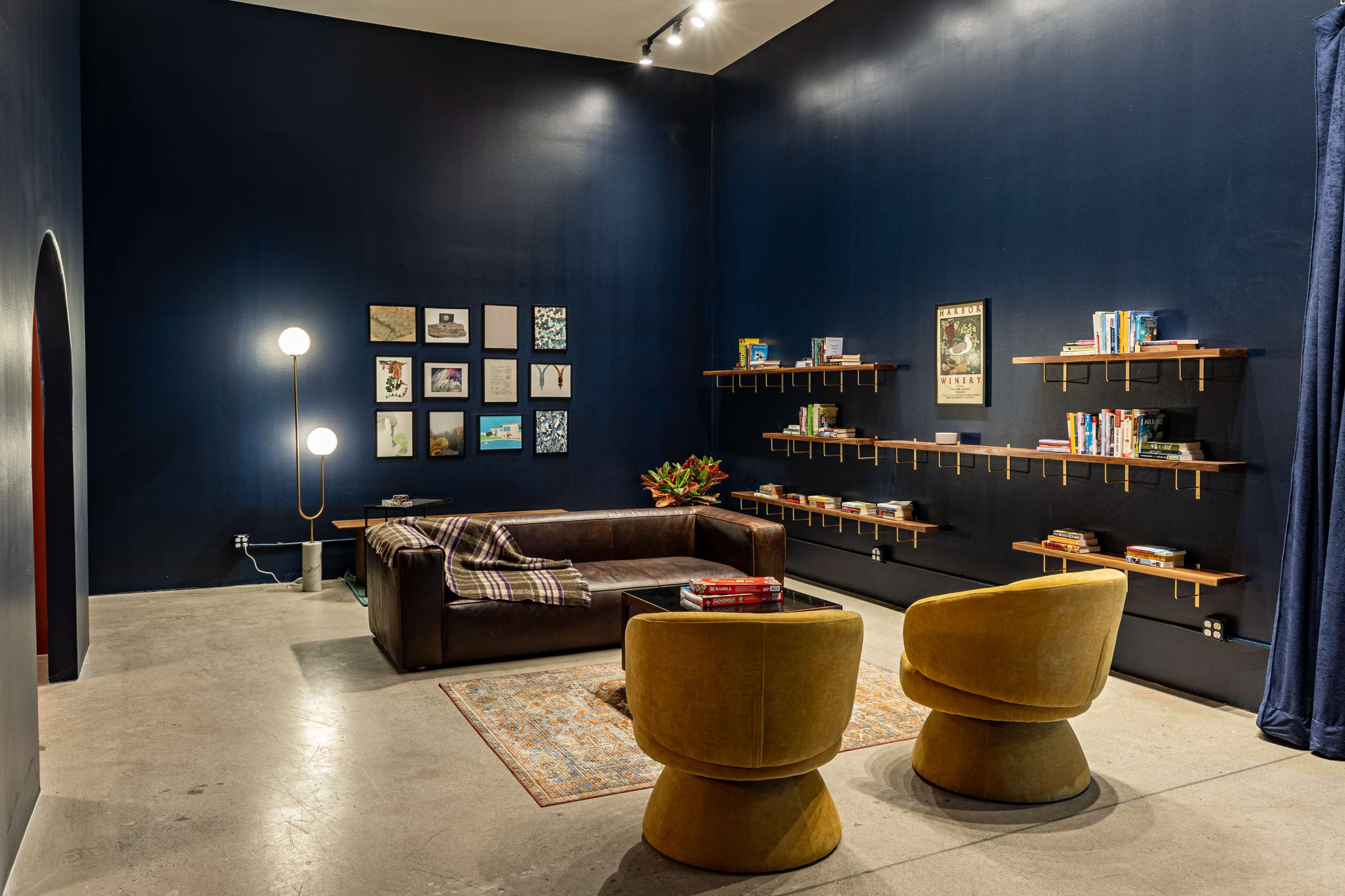A modern lounge area features a dark blue wall, a brown sectional couch, two yellow chairs, a patterned rug, and wall-mounted shelves displaying books and artwork.