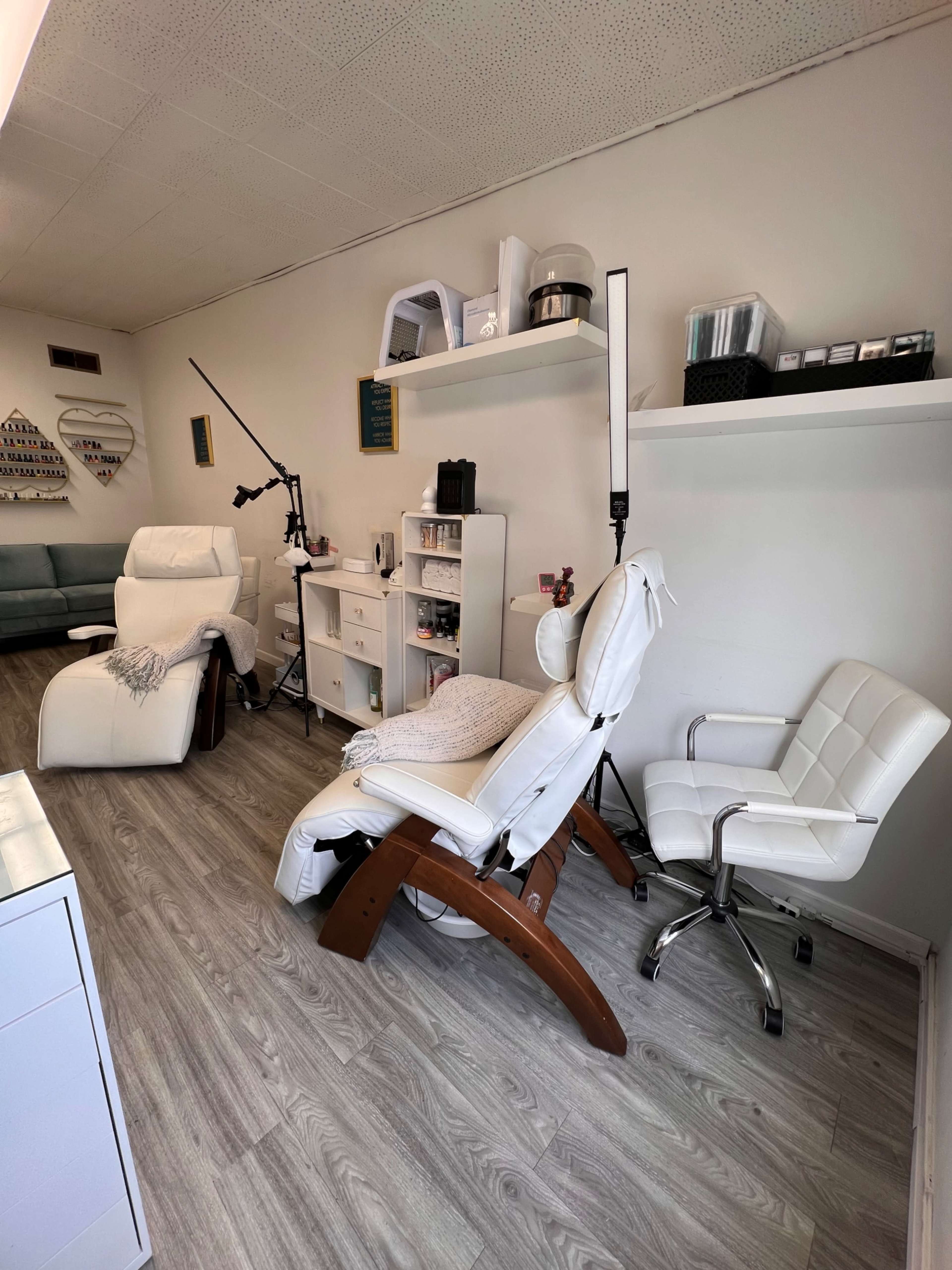 The image shows a small, well-organized beauty treatment room with two reclined chairs, a white rolling chair, and various equipment on shelves.