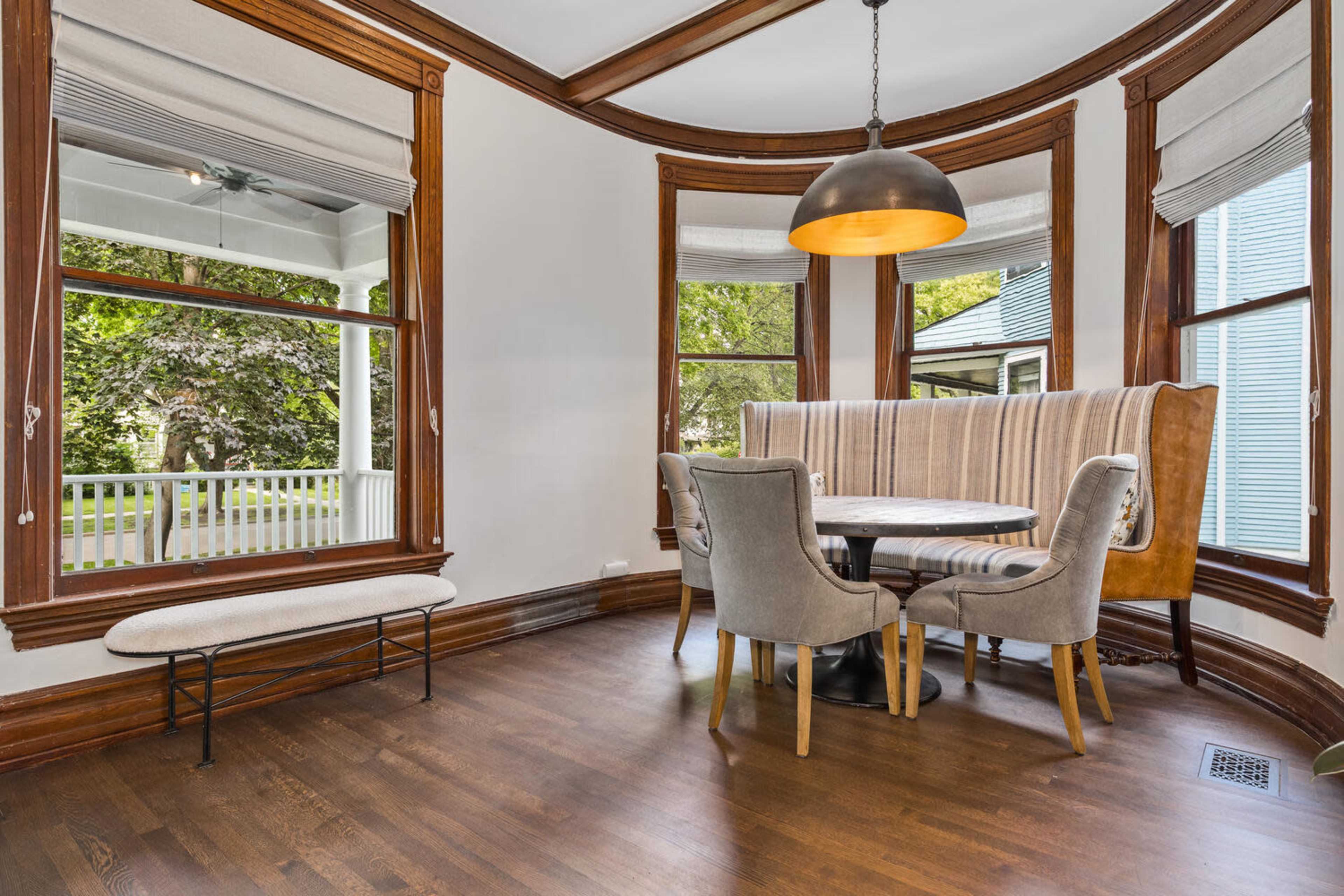 A round dining table with upholstered chairs is set in a bay window area, accompanied by a bench along the wall.