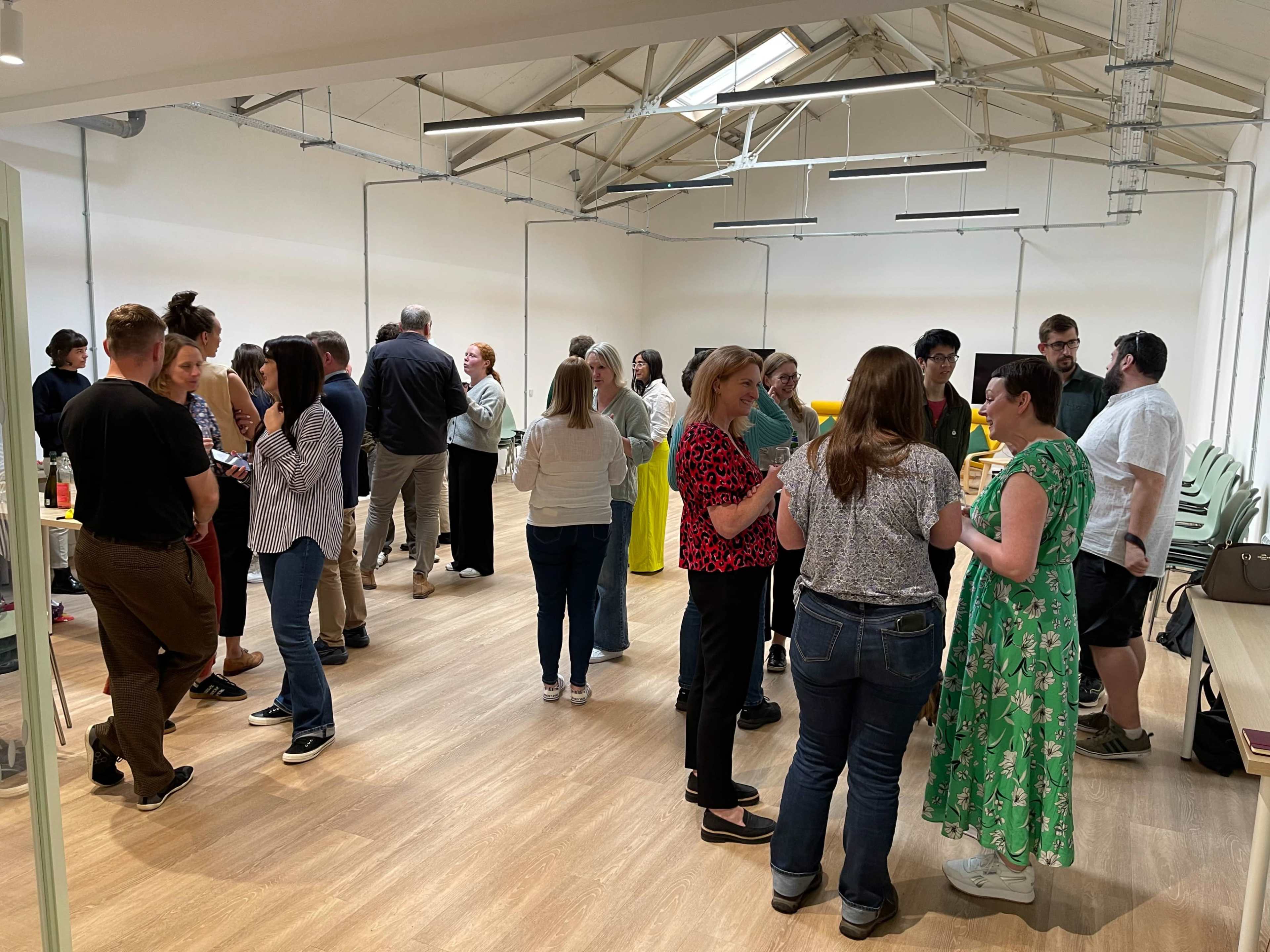 A group of people are mingling in a spacious room with bright lighting and a wooden floor.
