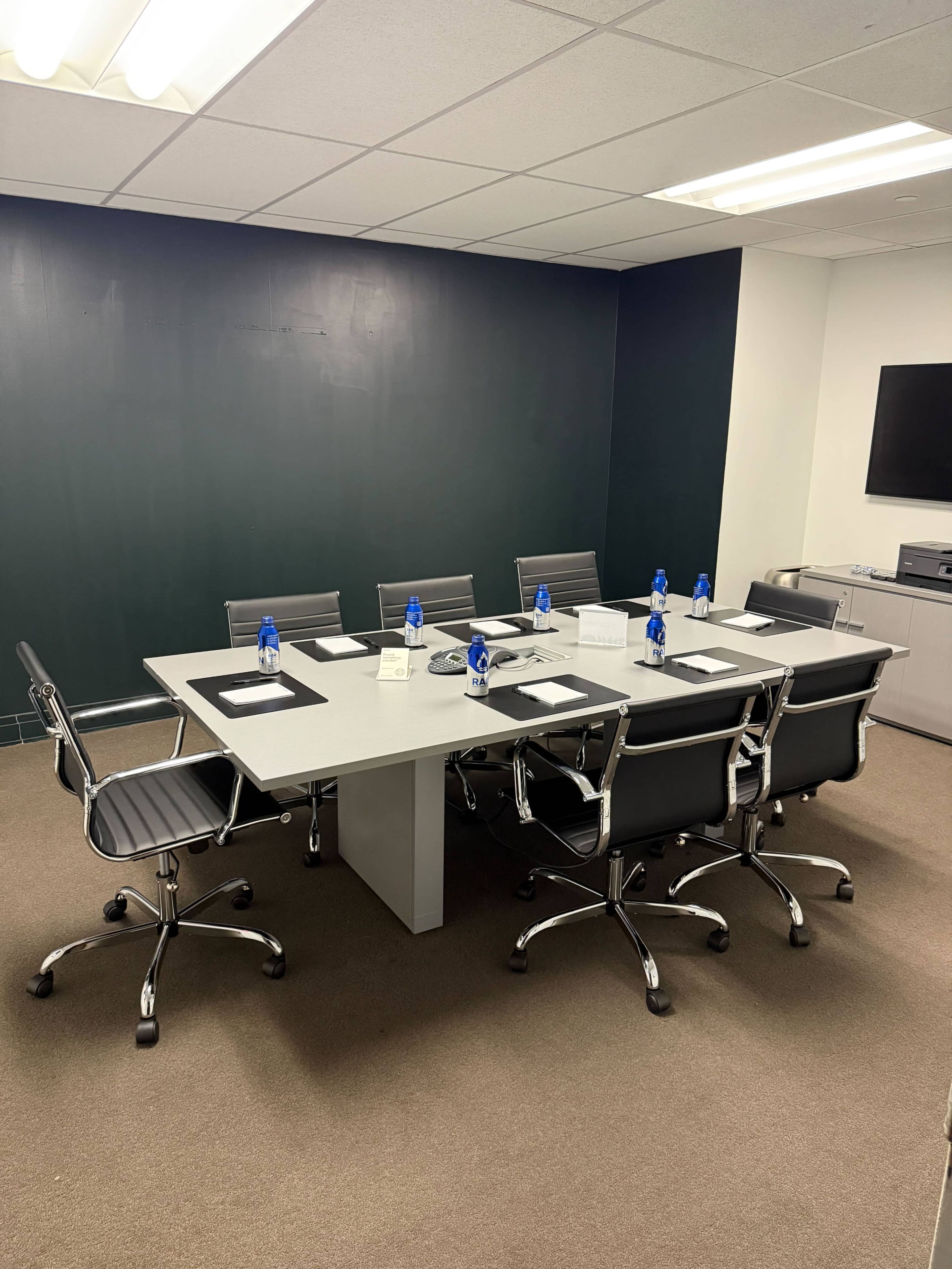 A conference room features a large rectangular table surrounded by ergonomic chairs, with water bottles and notepads placed at each seat.