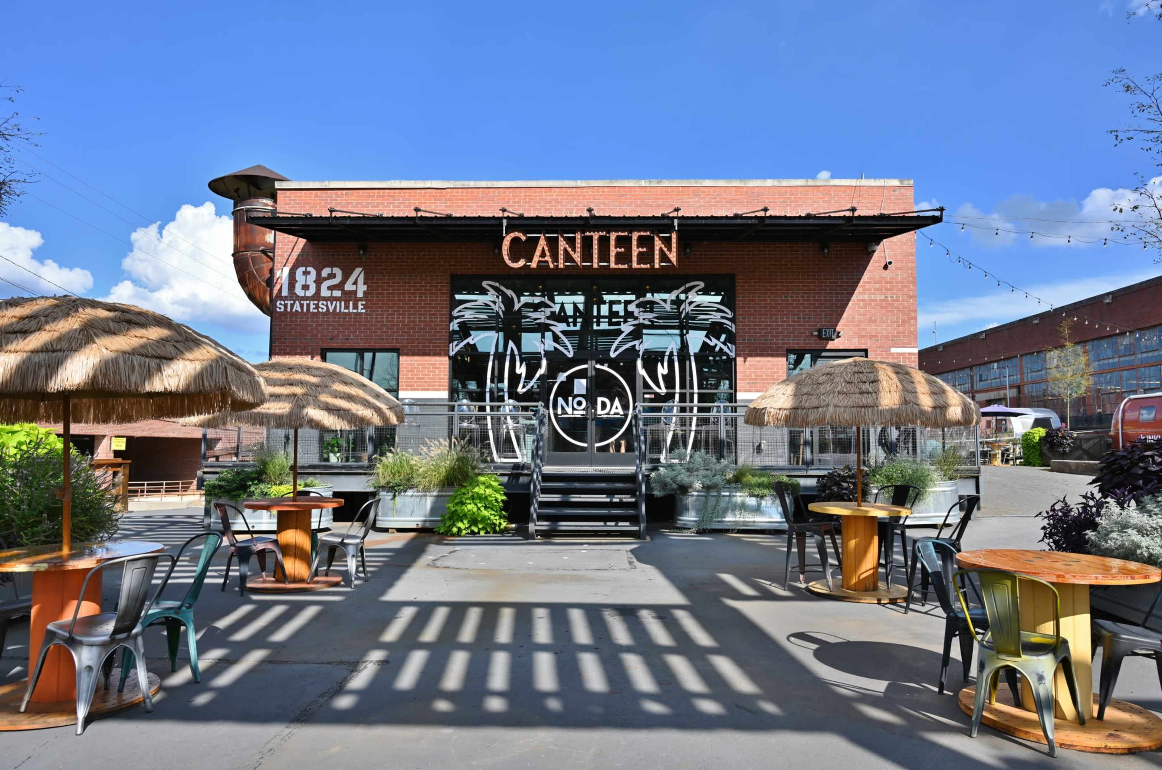 A vibrant outdoor dining area in front of a brick restaurant called "Canteen," featuring thatched umbrellas and colorful tables.