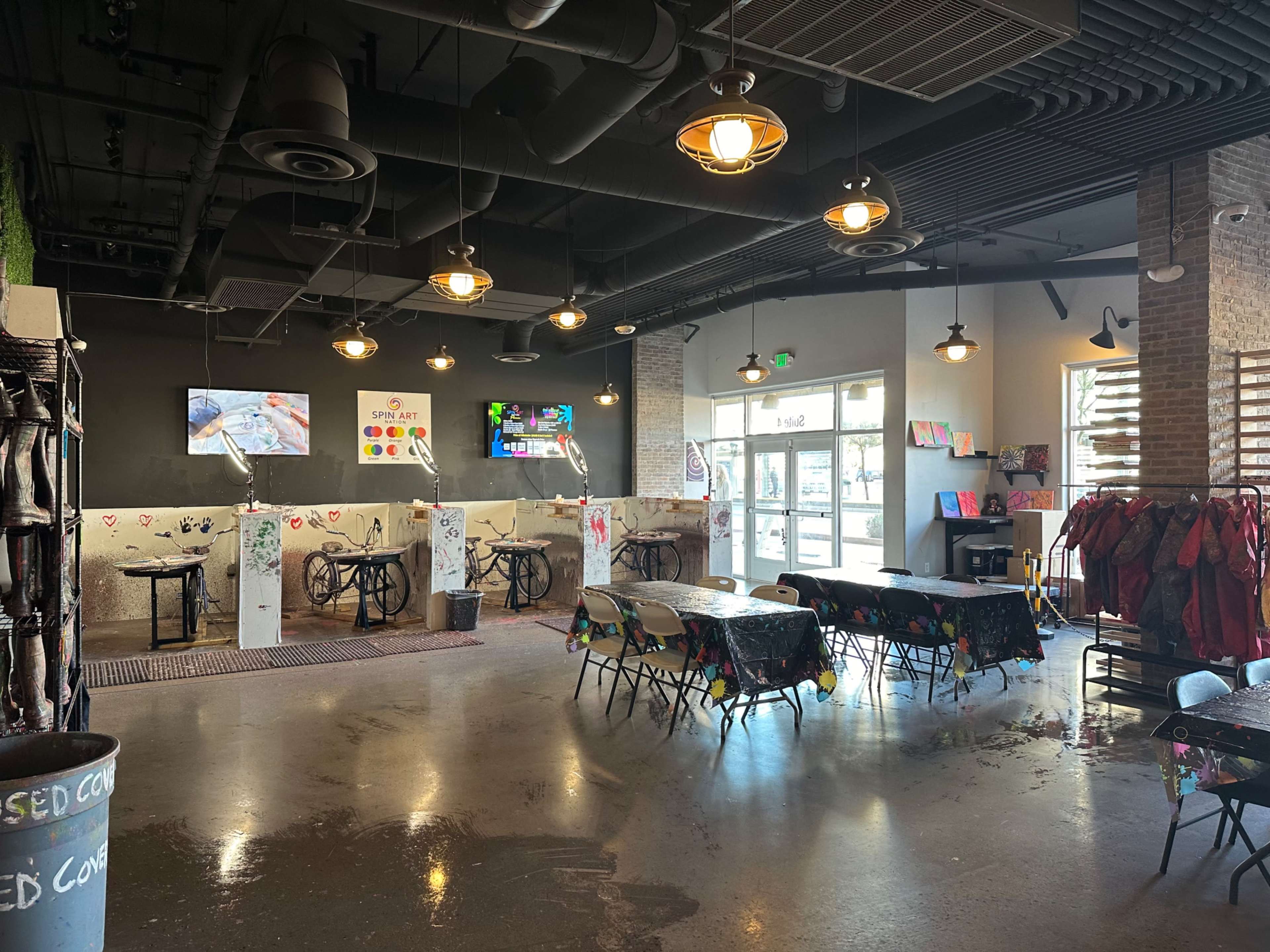 An indoor art studio with tables covered in paint splatters, several easels set up for painting, and light fixtures hanging from the ceiling.