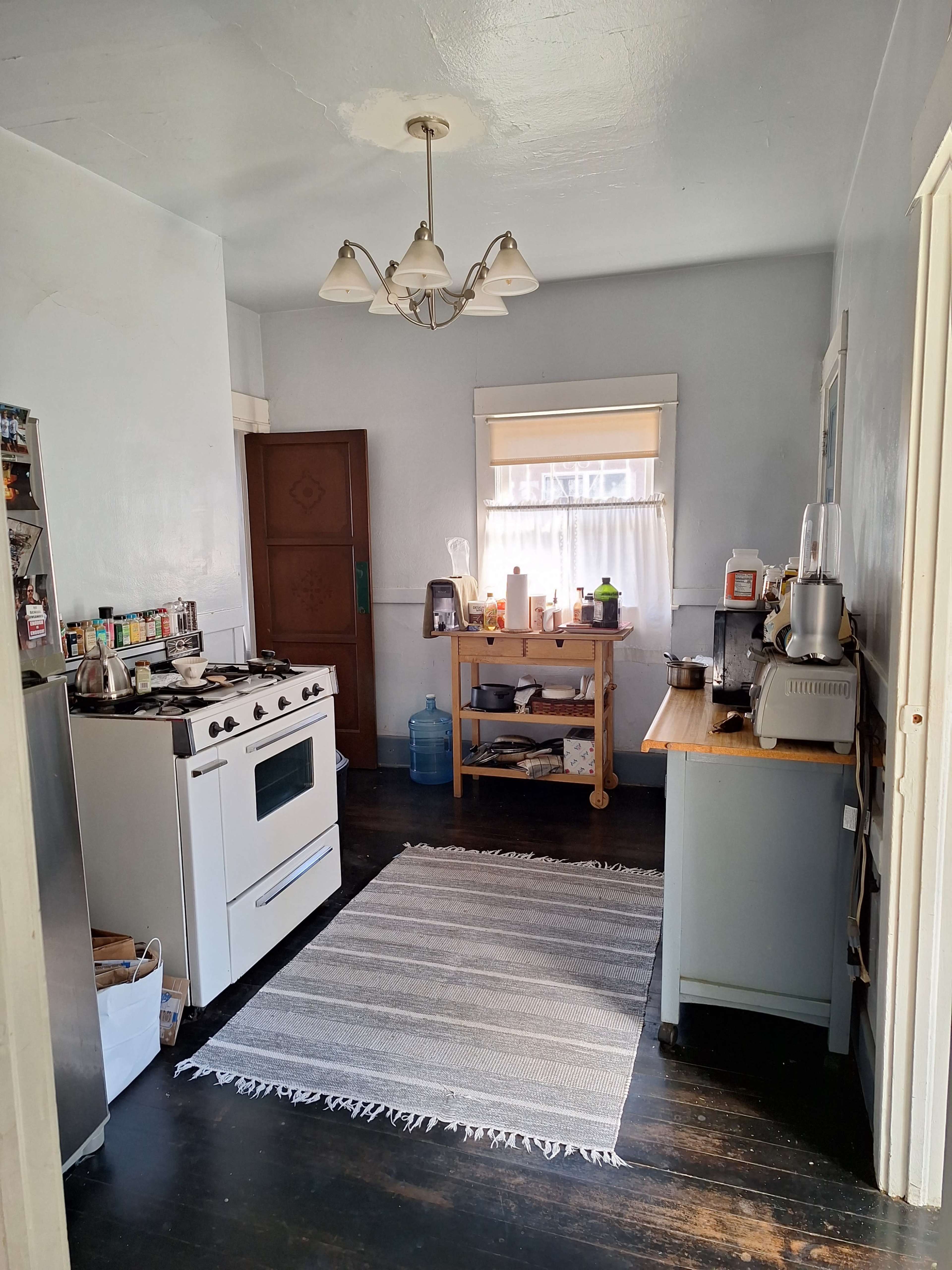 The image depicts a small, well-lit kitchen featuring a white stove, a wooden preparation table, and a striped rug on the dark wooden floor.