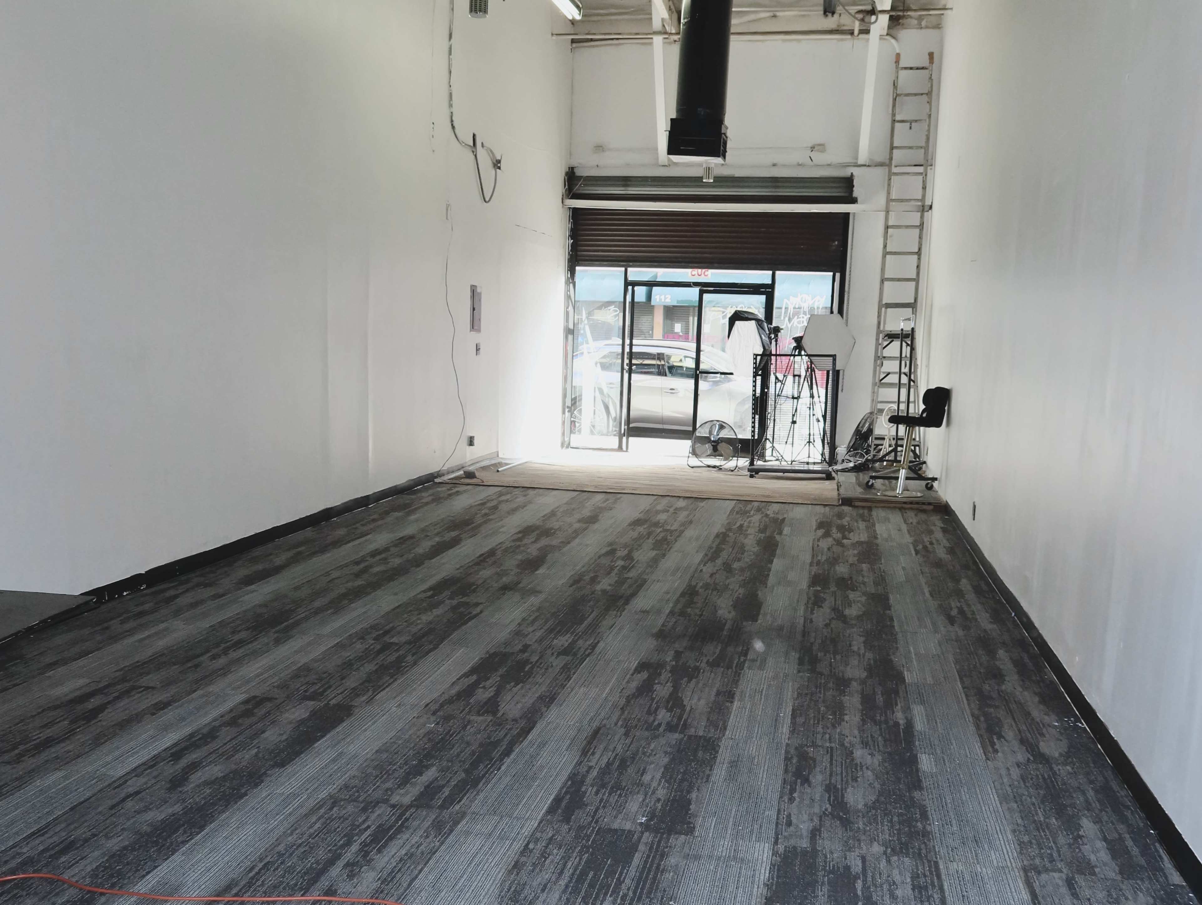 The image shows an empty retail space with a bare floor, a closed window shutter, and a ladder against the wall.